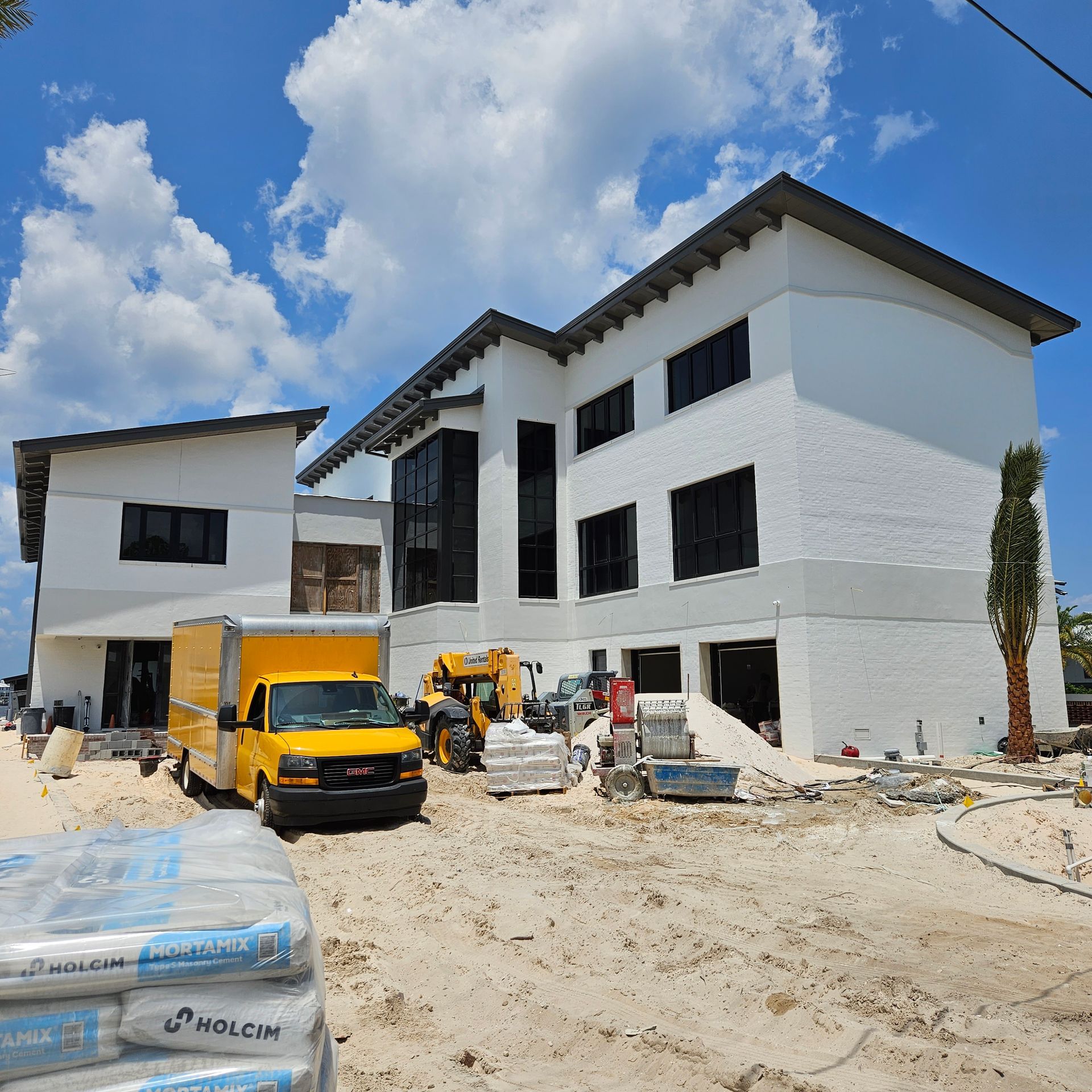 A yellow delivery truck is parked at a construction site in front of a white two-story building under a blue sky.