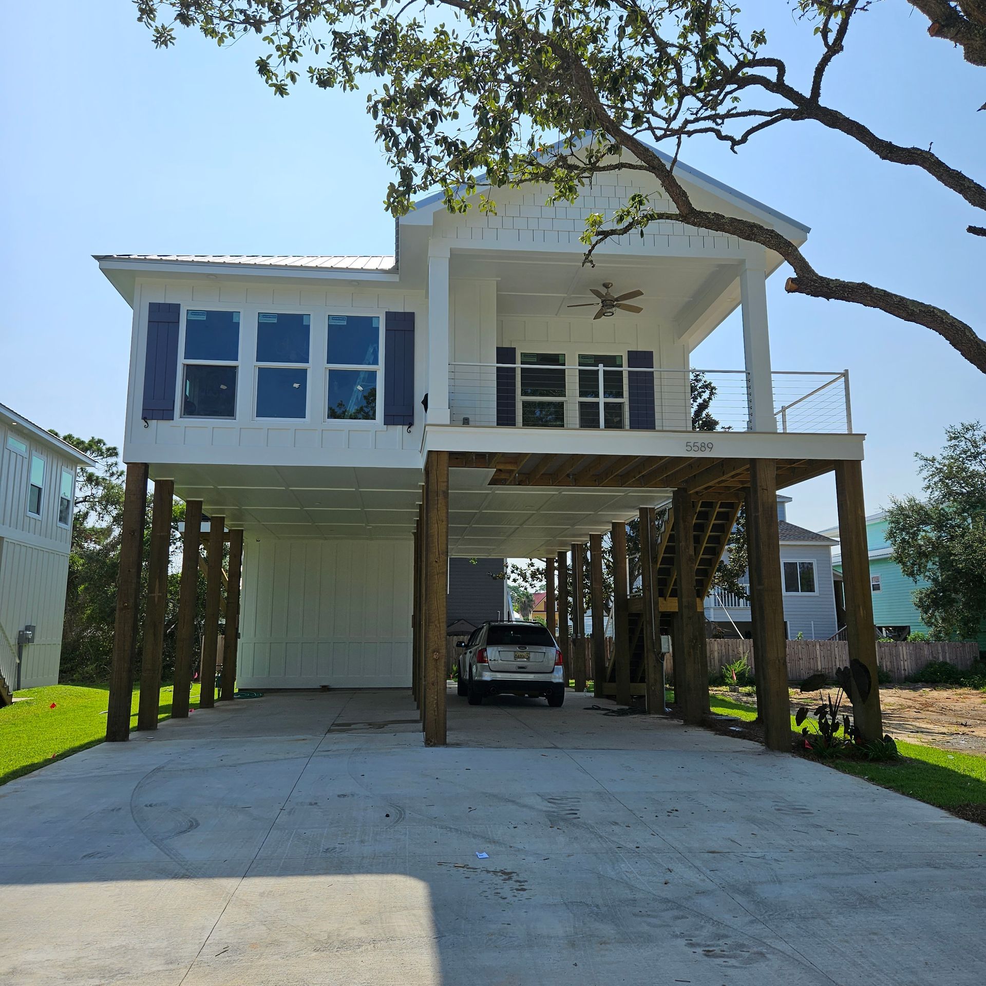 A two-story coastal home on tall wooden pilings with a white exterior, blue shutters, and a front porch under a clear sky.