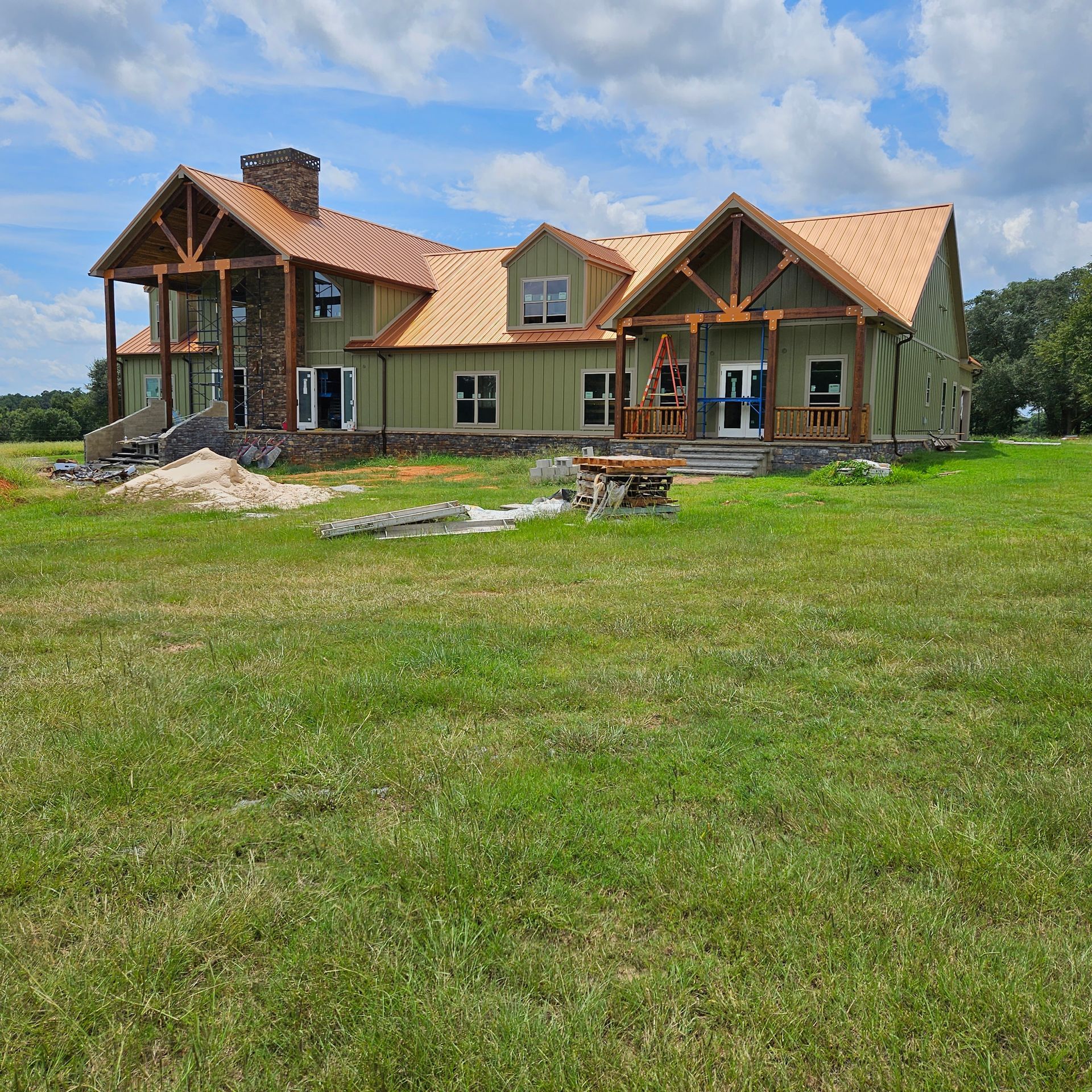 A large, two-story house under construction with a metal roof and green siding, set in a sunny grassy field.