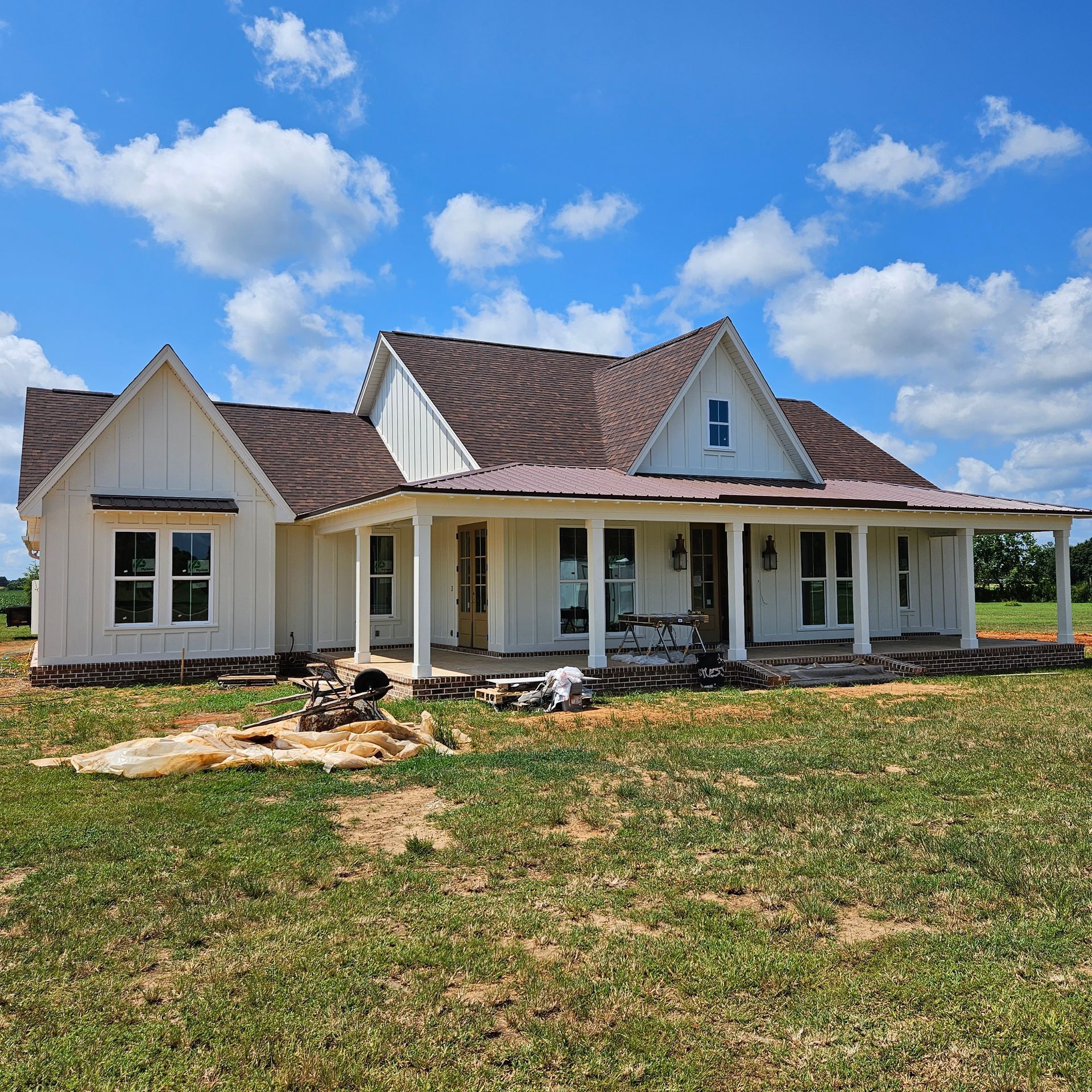 A newly constructed white farmhouse with a brown metal roof, a front porch, and tall windows under a bright blue sky.