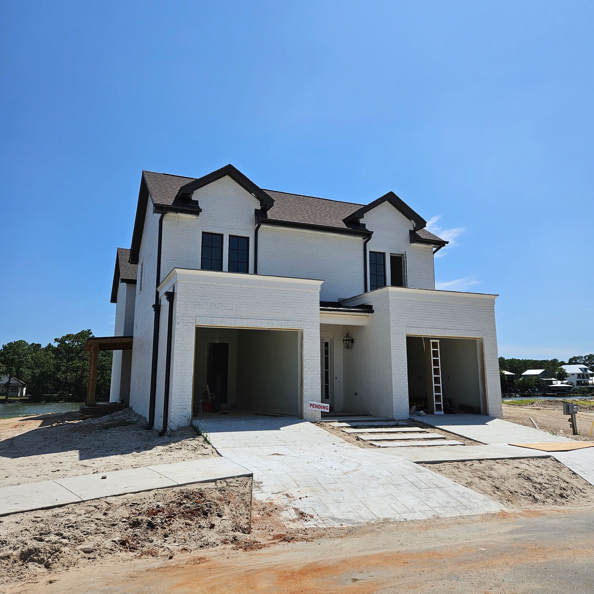 A two-story white duplex under construction with two open garage bays, dark roof gables, and a dirt-covered yard.