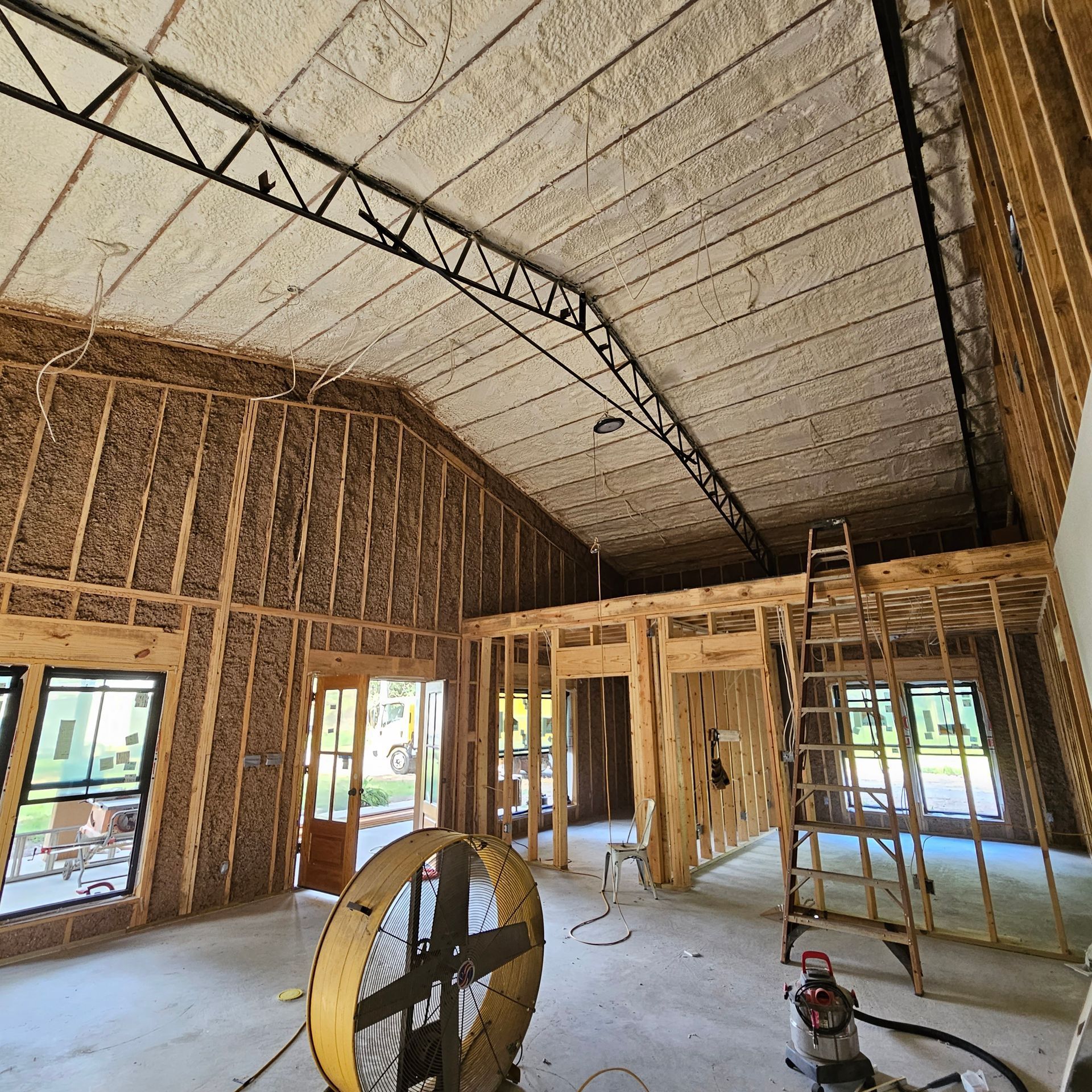 The interior of a construction site showing wood-framed walls, a high vaulted ceiling, a metal ladder, and a large fan.