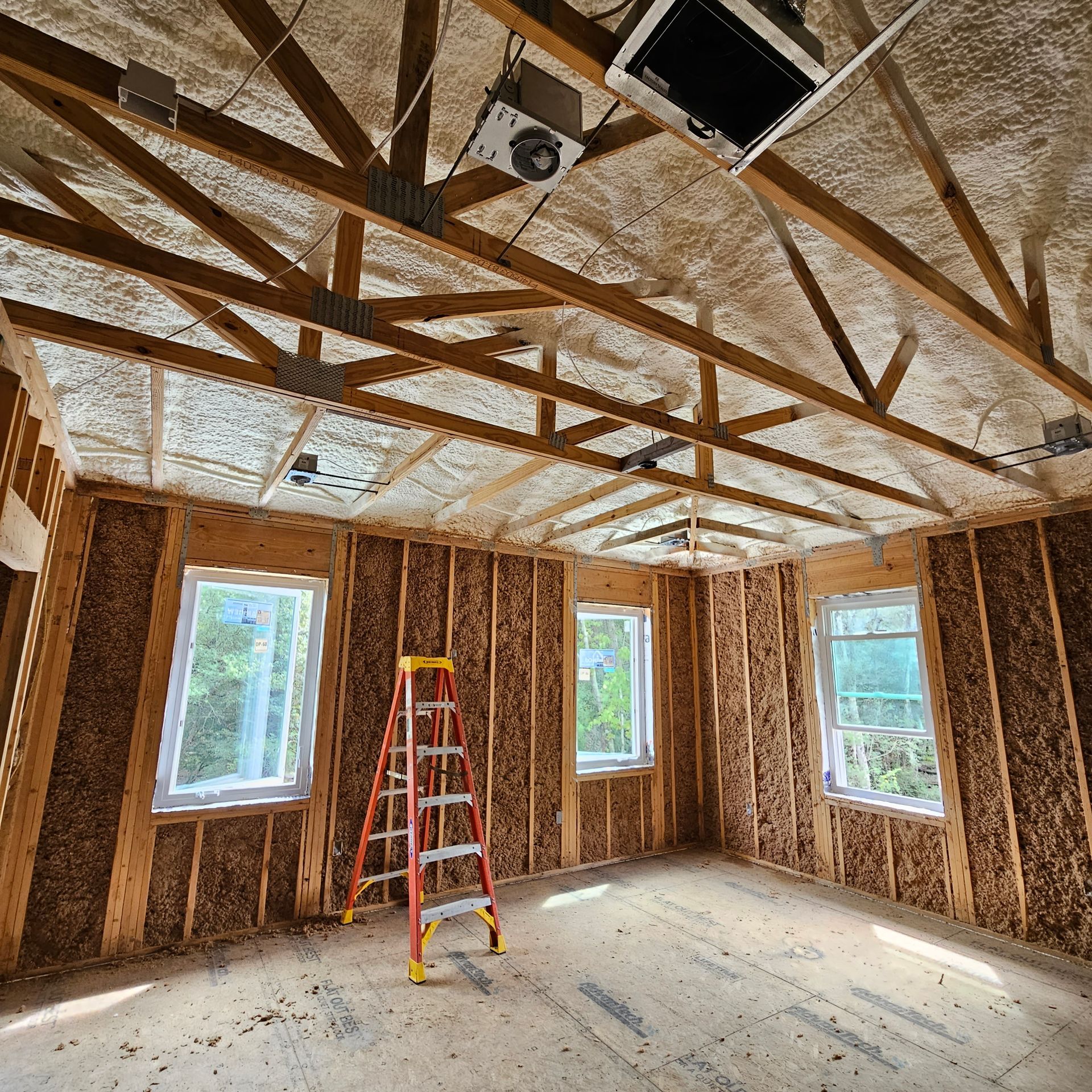 An unfinished room under construction, featuring exposed wooden roof trusses, insulated walls, and a central red ladder.