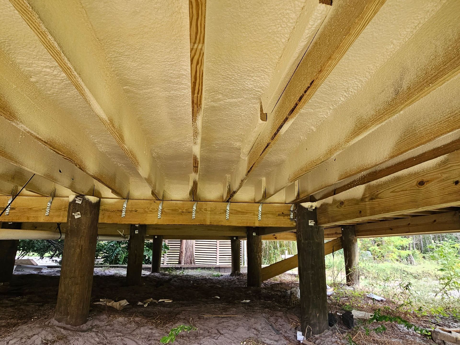 The underside of a wooden deck with yellow spray foam insulation applied between the floor joists, supported by posts.