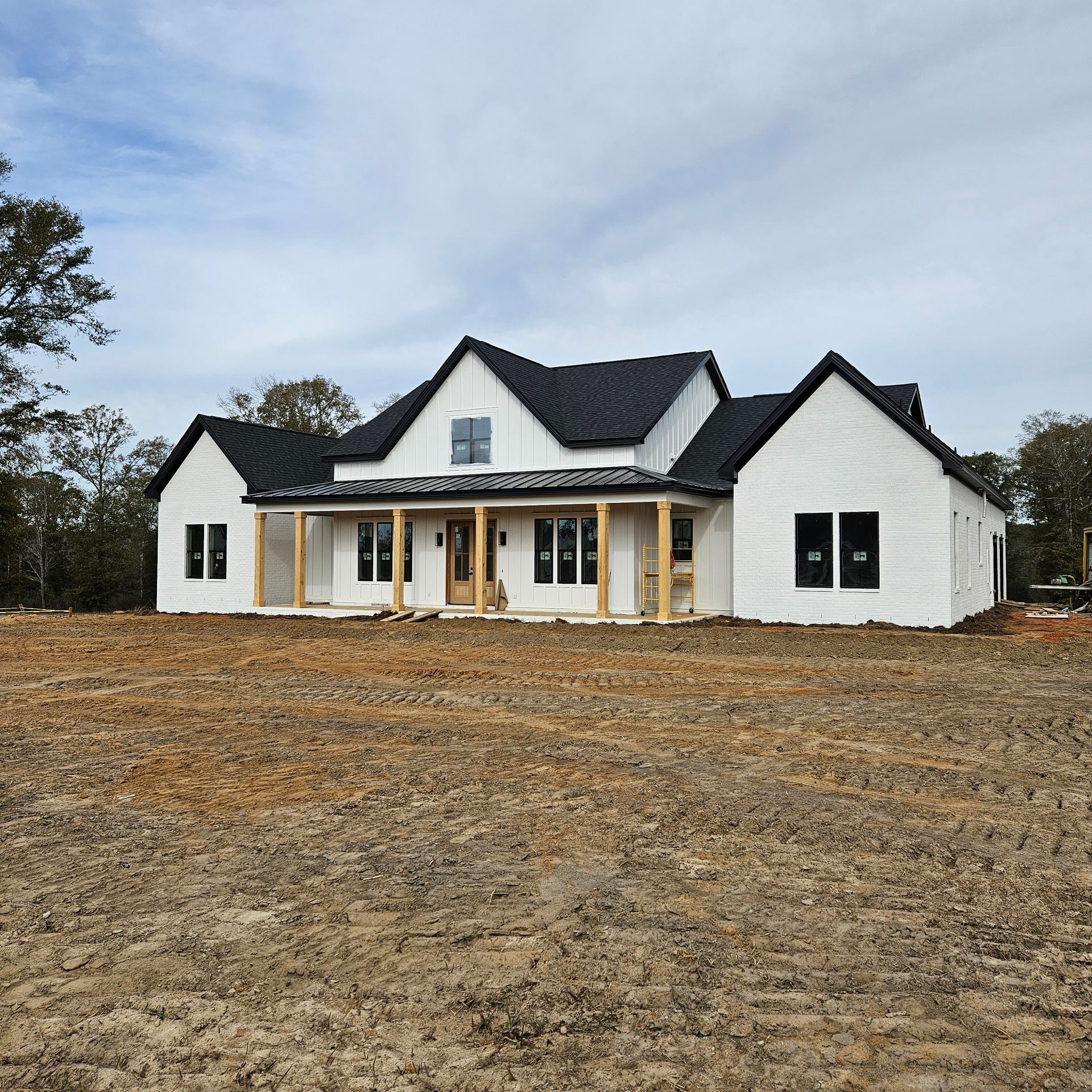 A newly constructed white brick house with black roofing and wooden porch posts sits on an unlandscaped dirt lot.