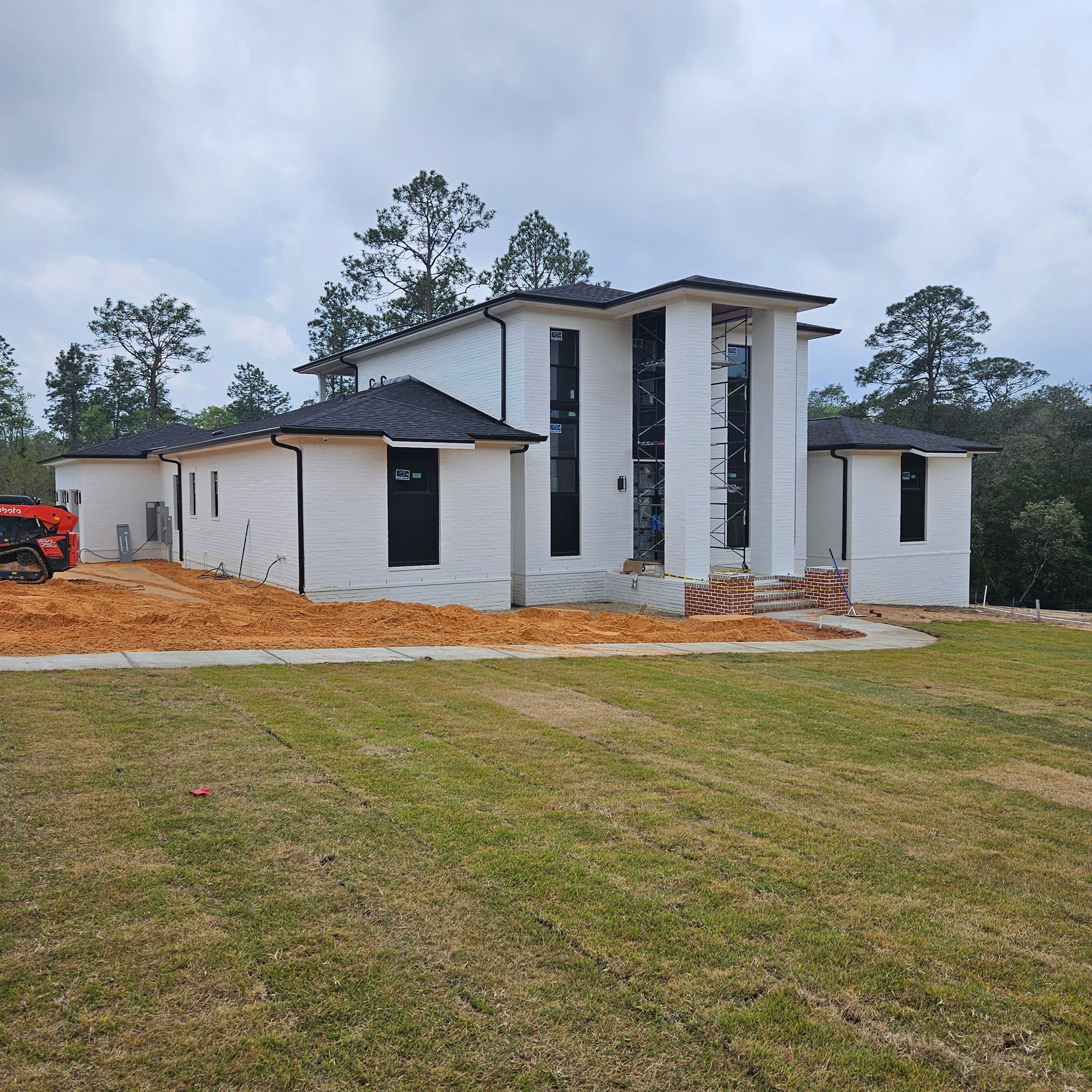 A modern white house under construction with black window frames and roof, set on a rural plot with dirt and grass.