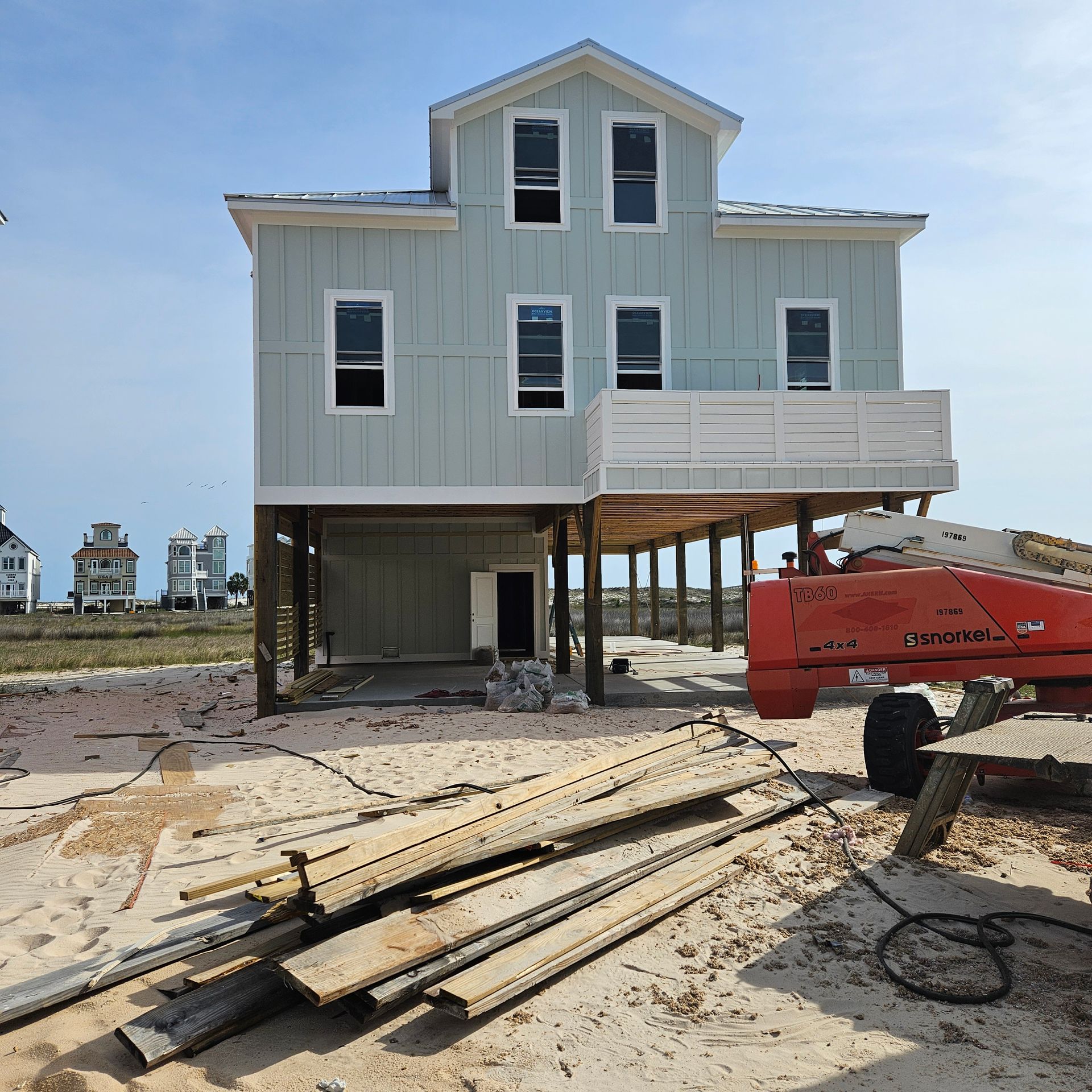 A light green elevated house under construction on a sandy lot with a red construction lift in the foreground.