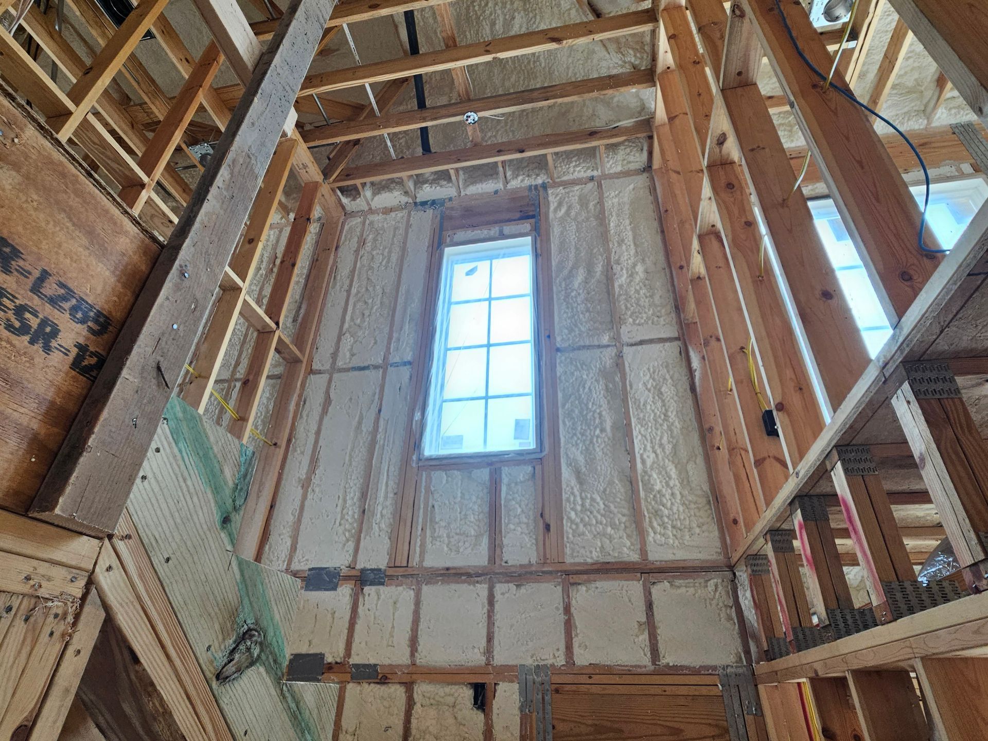 Interior view of a residential construction site featuring tall wooden framing, spray foam insulation, and a window.