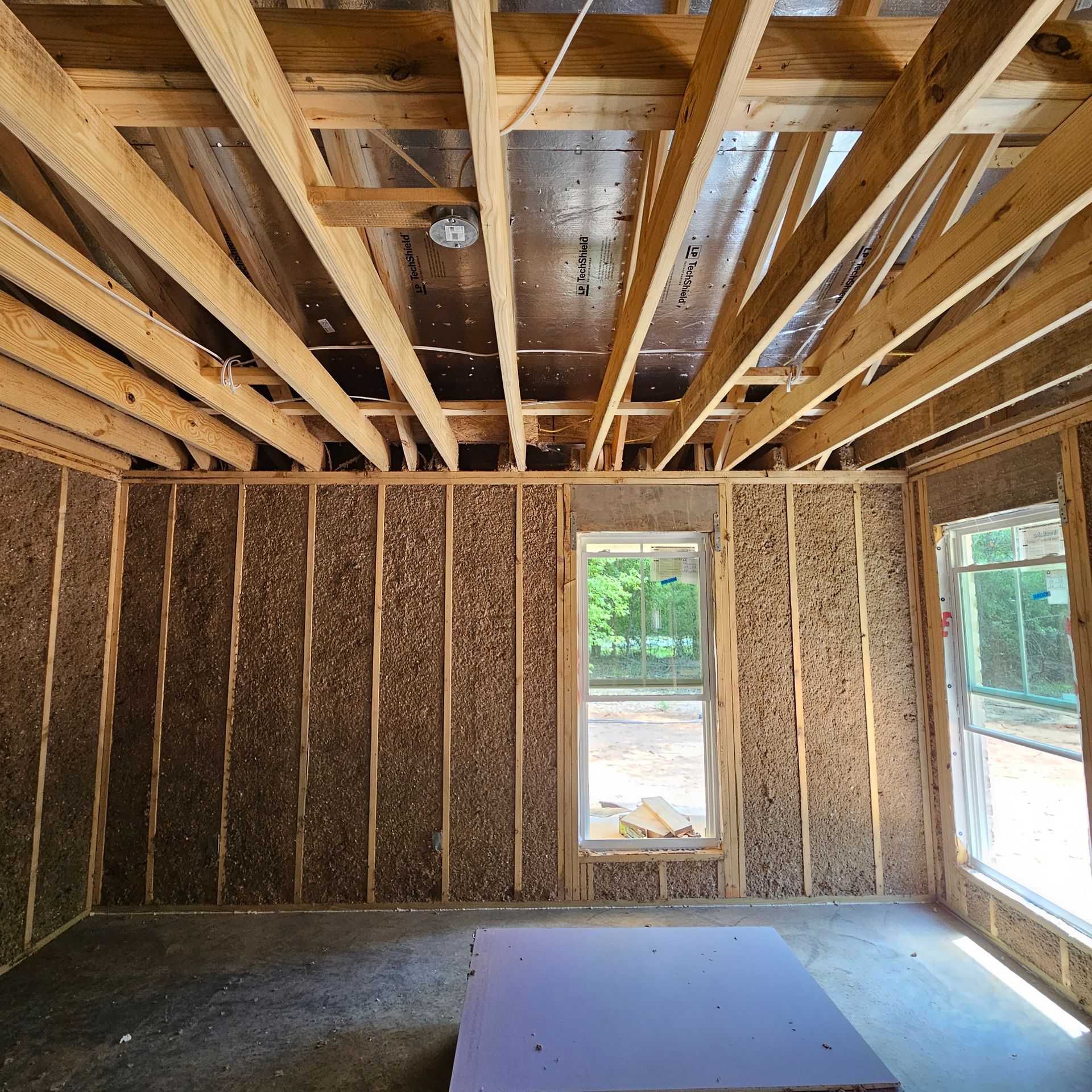 Interior view of a residential room under construction with wood wall studs, exposed rafters, and two installed windows.