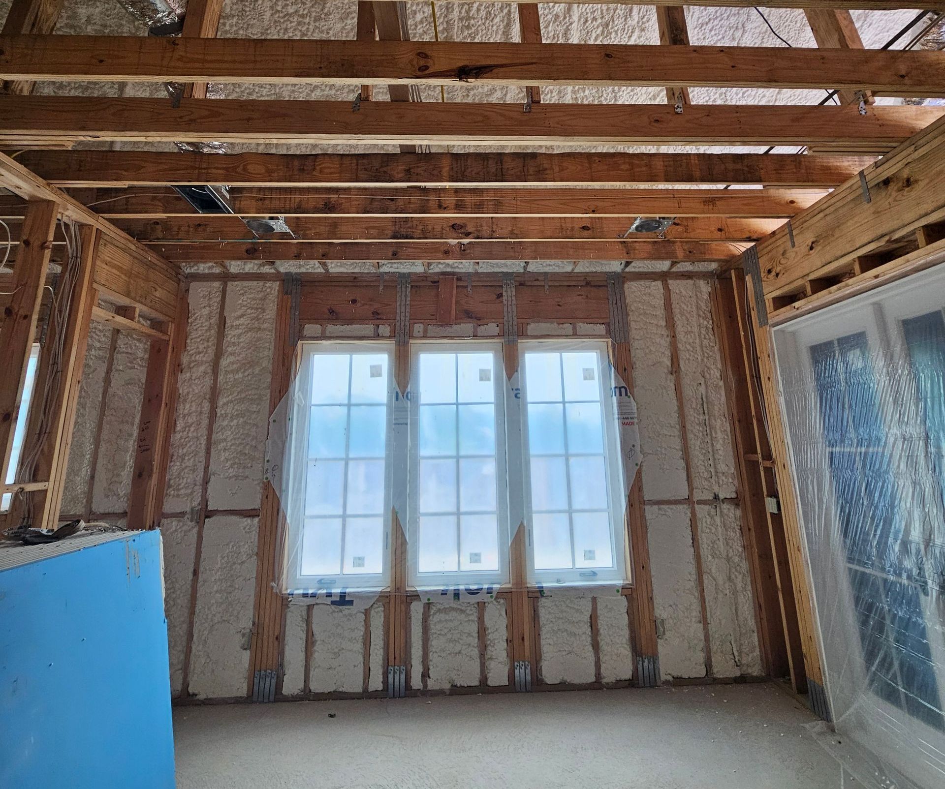 An unfinished room featuring bare wood framing, white spray foam insulation, and three windows centered on the back wall.