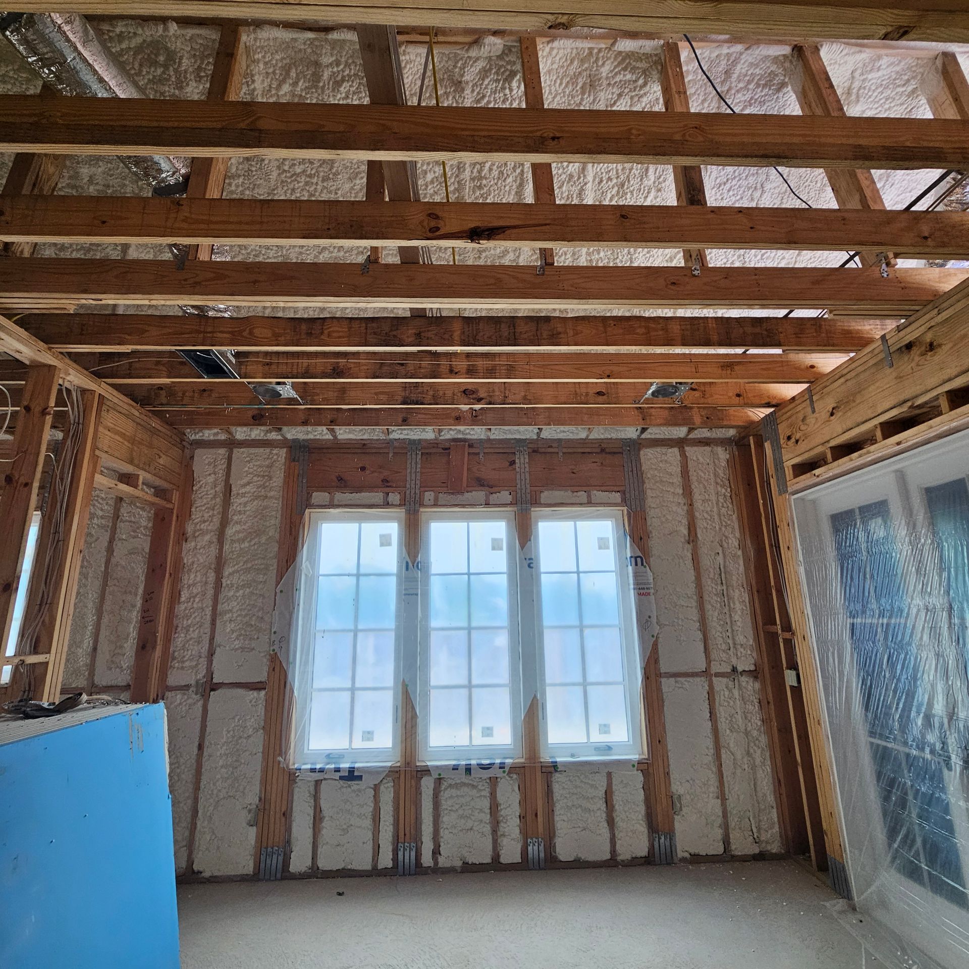 An unfinished room with exposed wooden ceiling joists and foam-insulated walls surrounding a three-pane window unit.
