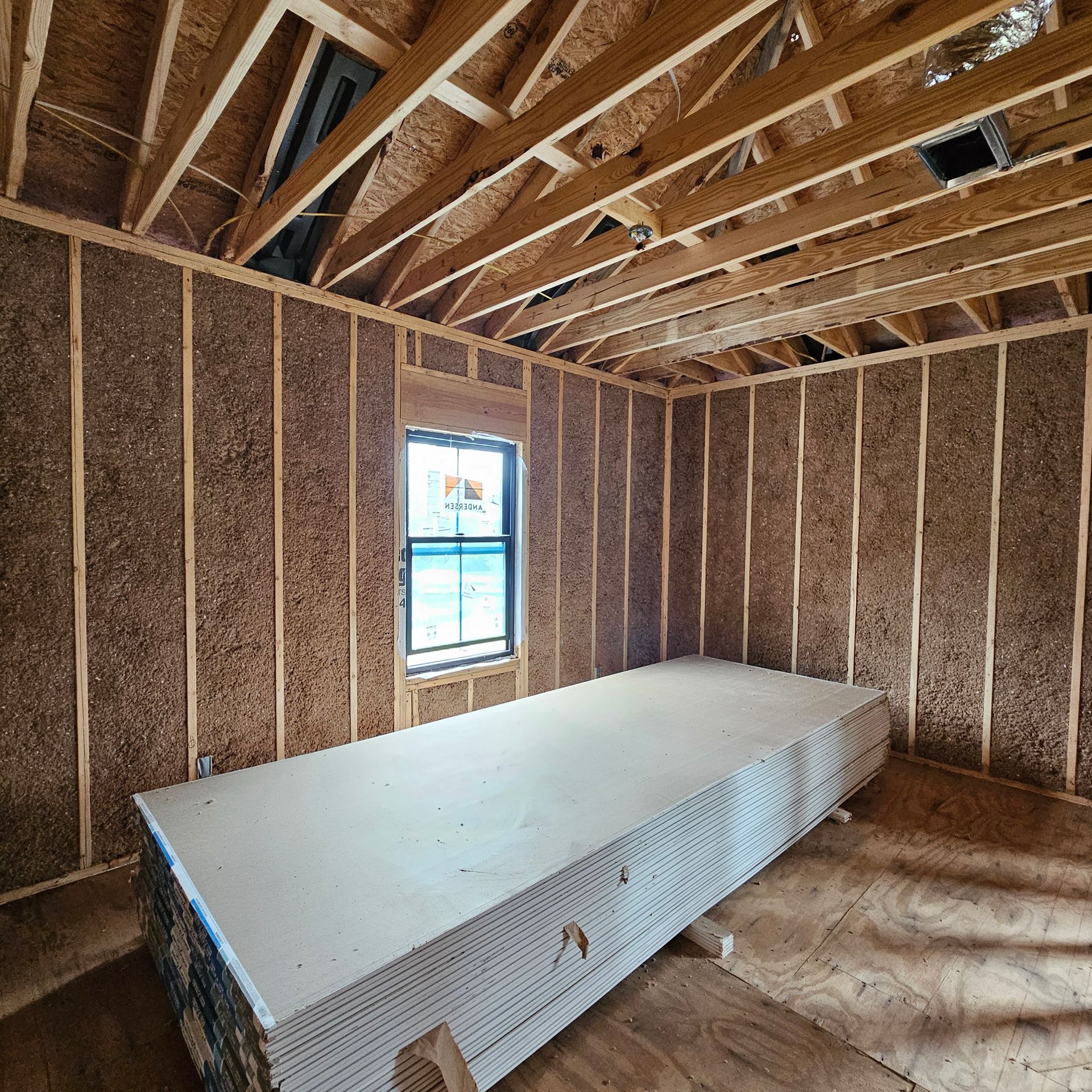 A stack of drywall panels sits in a room under construction with exposed wooden wall studs and roof rafters and a window.
