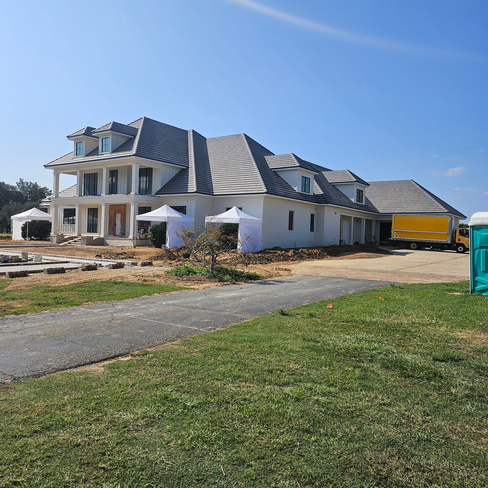 A large, multi-story white house under construction with a grey tiled roof, surrounded by dirt, tents, and a yellow truck.