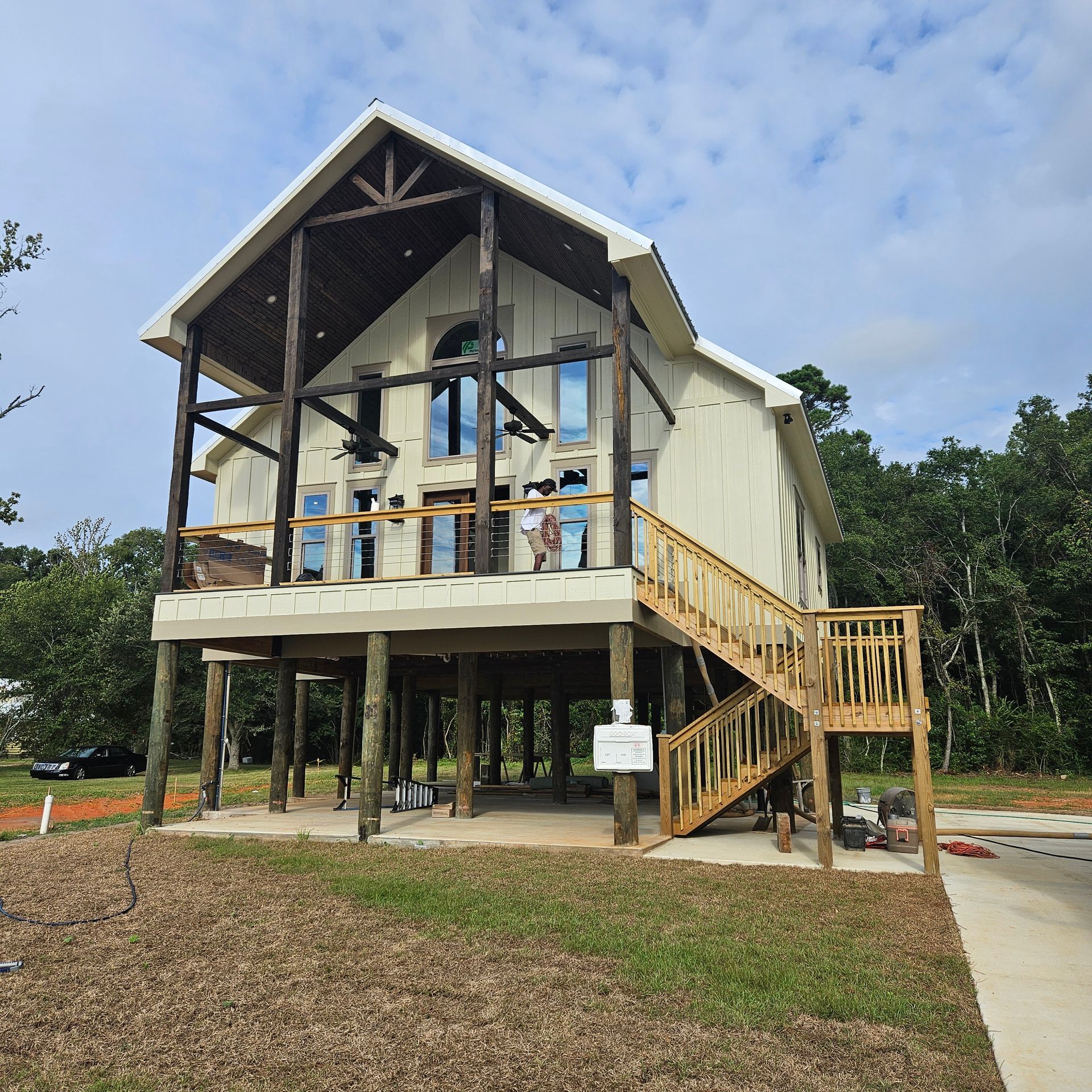 A modern, elevated wooden house on stilts with a deck and staircase, surrounded by greenery under a cloudy sky.