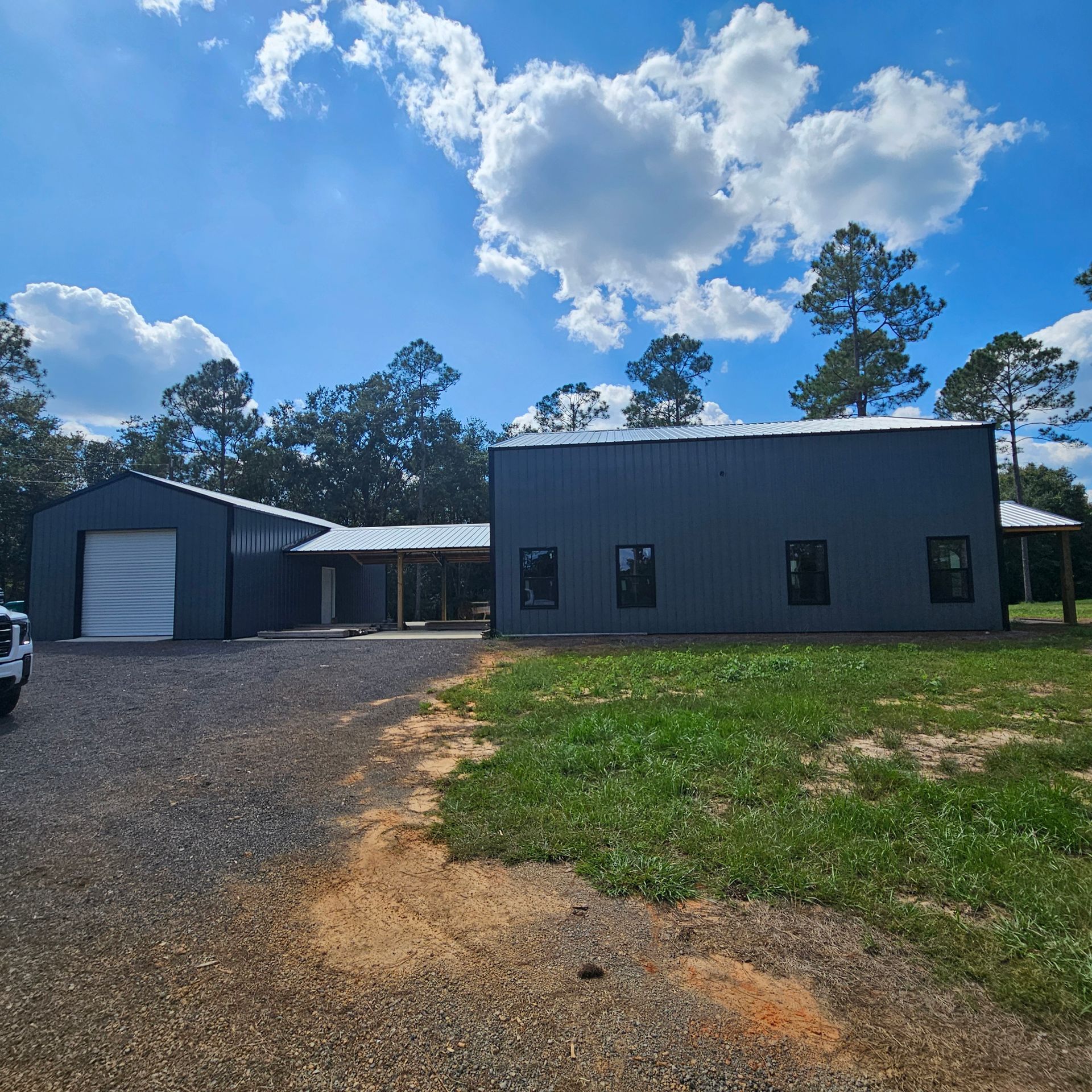 A dark grey metal barn-style building with a white garage door, connected by a covered walkway under a bright blue sky.