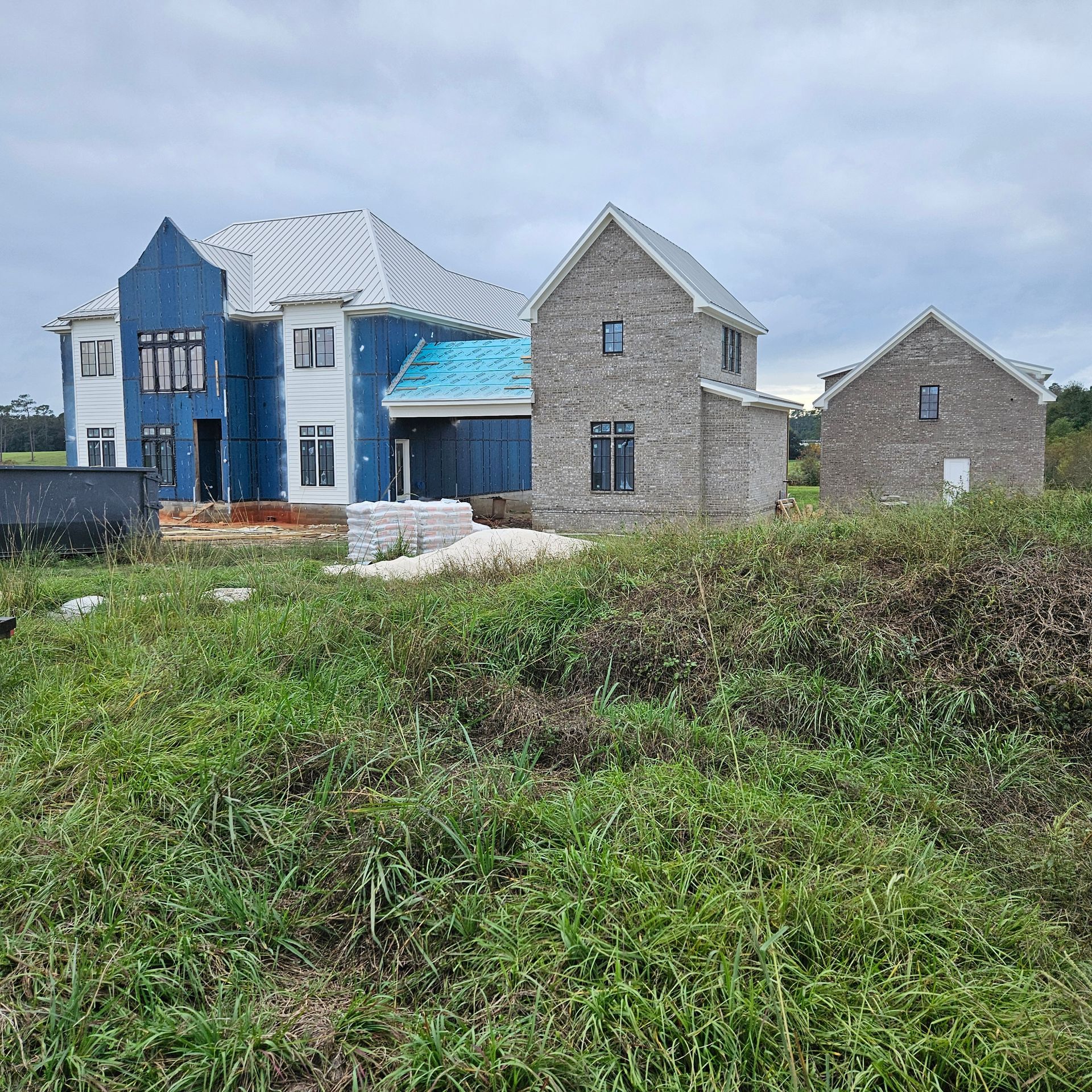 Three houses in various stages of construction stand in a grassy field under a cloudy sky.
