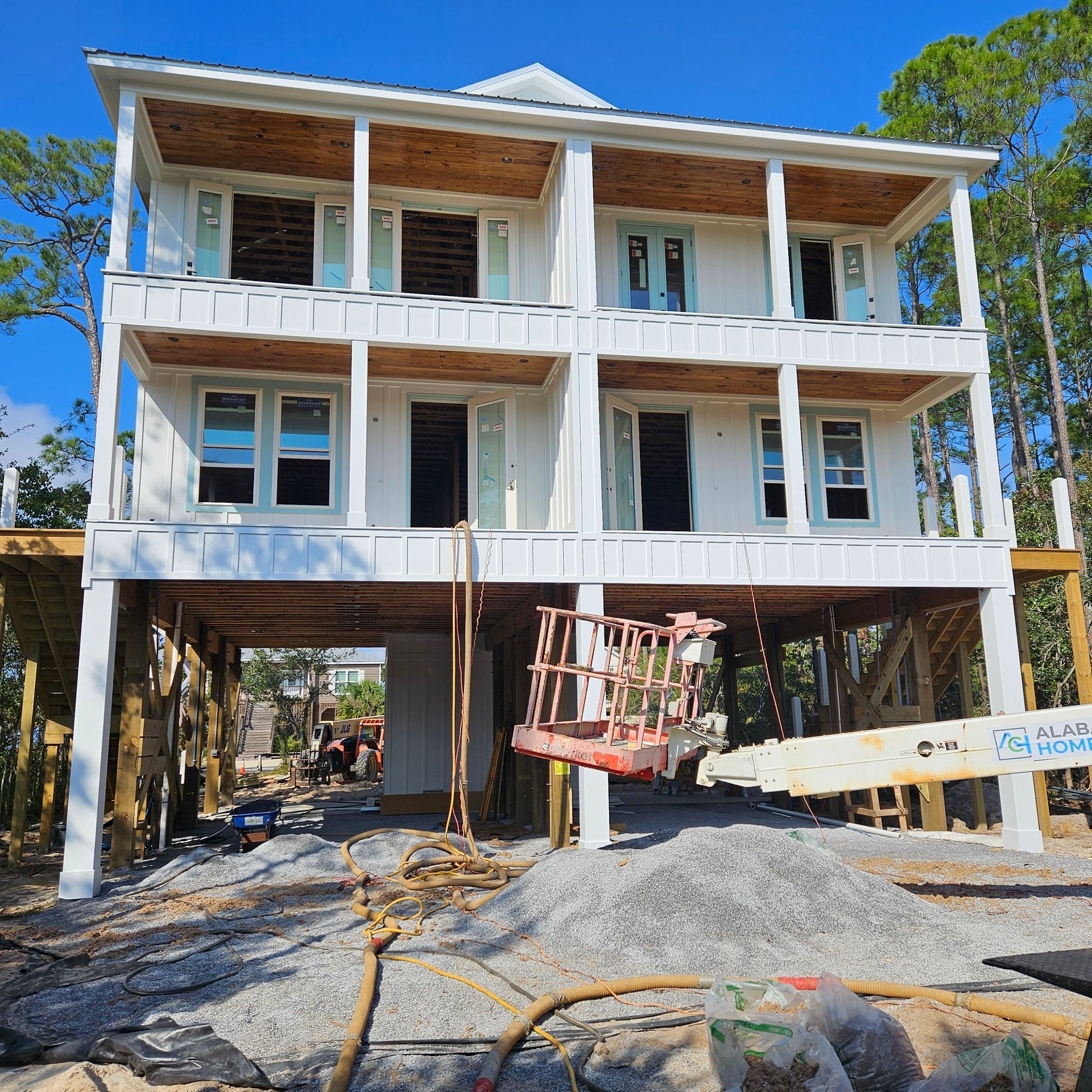 A three-story white house under construction on stilts, with a crane basket in front and gravel on the ground.