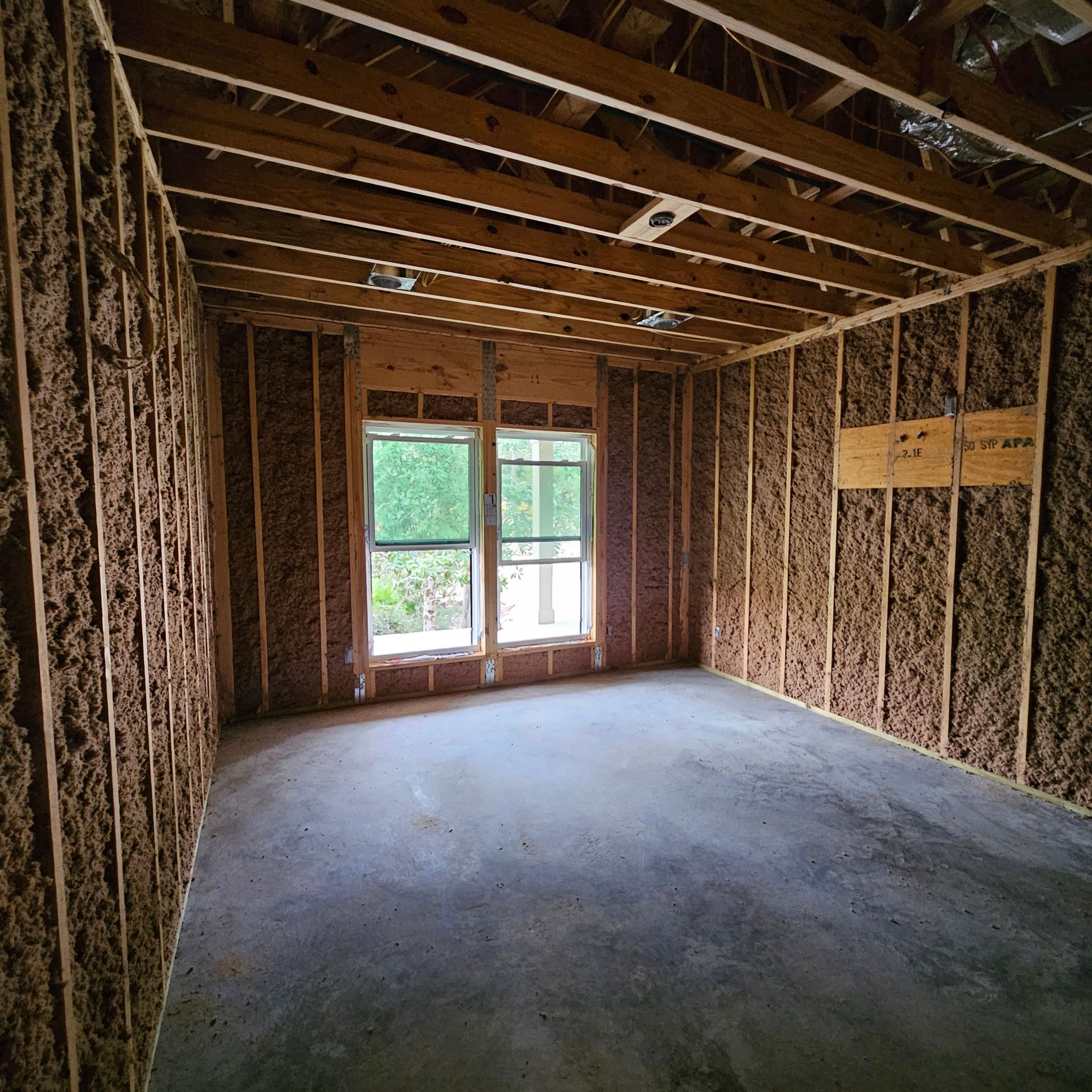 Interior of a room under construction with exposed wooden framing, brown insulation between wall studs, and concrete floor.