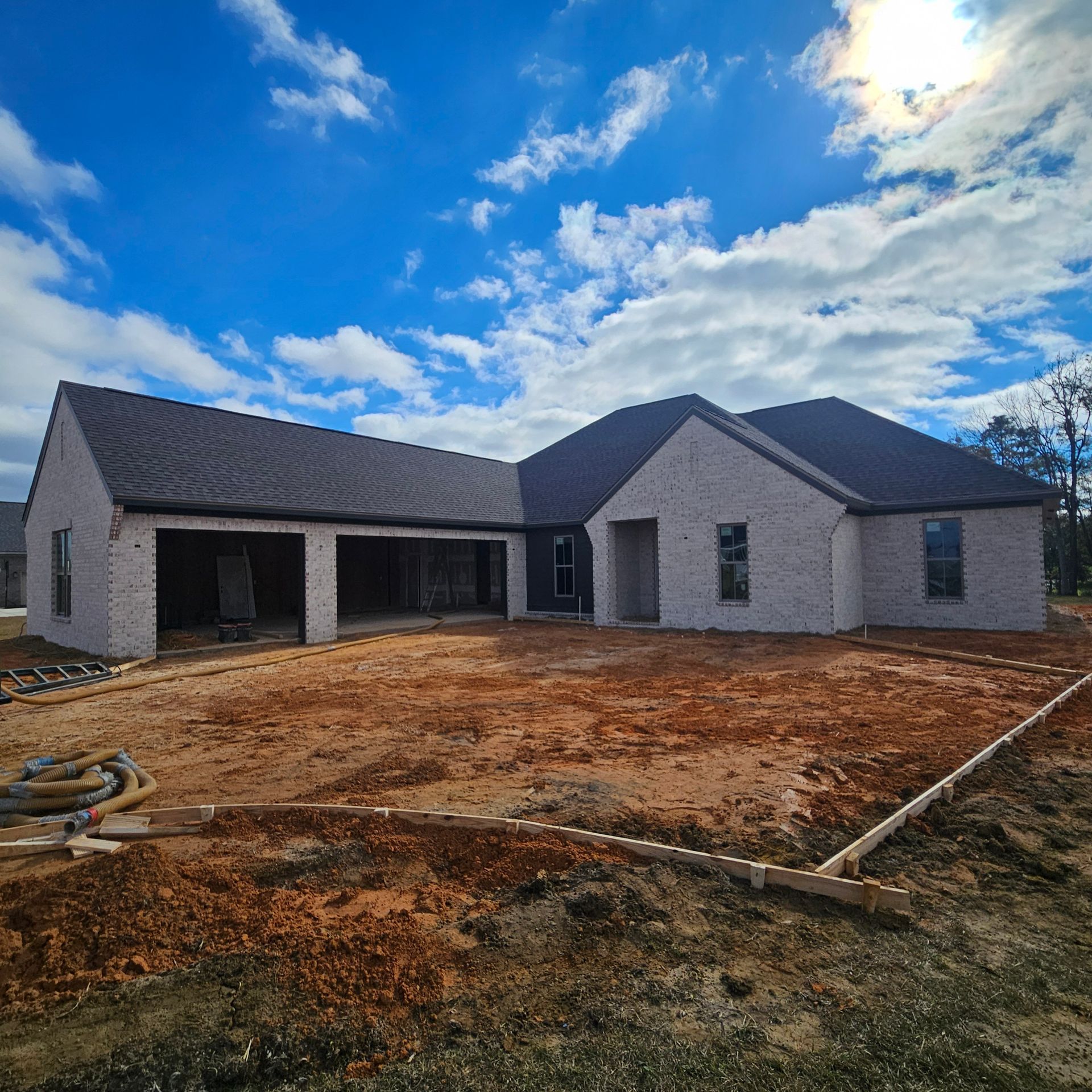 A single-story brick house under construction with a dark roof and open garage, set against a bright blue sky.