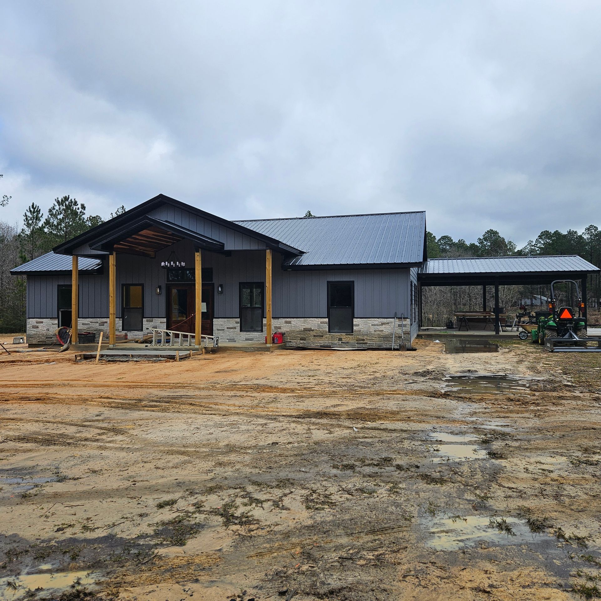 A modern, grey-sided single-story house with a metal roof and stone foundation sits on a muddy, unfinished construction site.