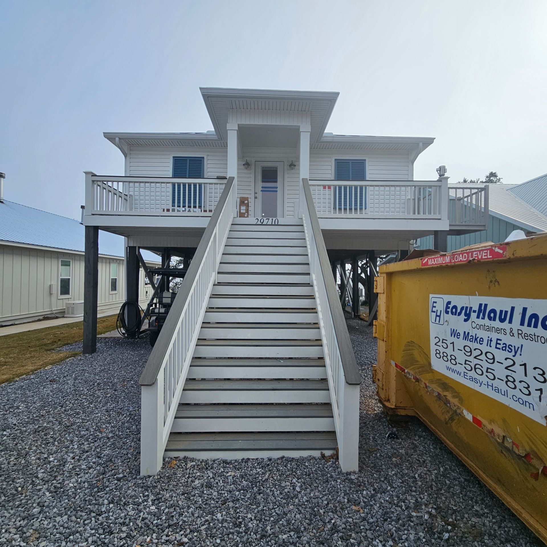 A white elevated stilt house with stairs leading to a front porch, featuring a yellow dumpster parked on a gravel lot.