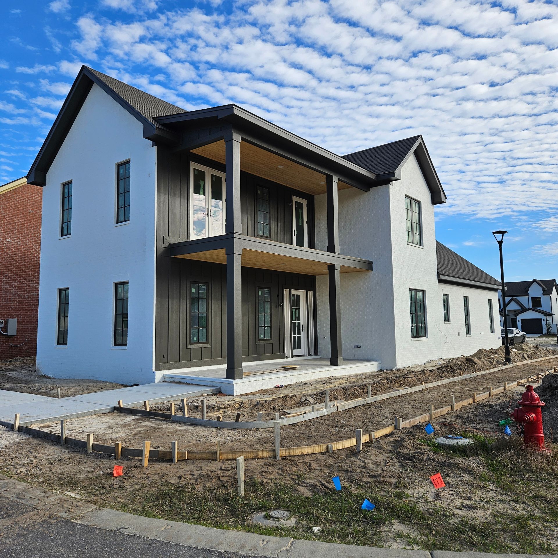 A two-story house under construction with white stucco, dark vertical wood siding, a covered porch, and blue sky.