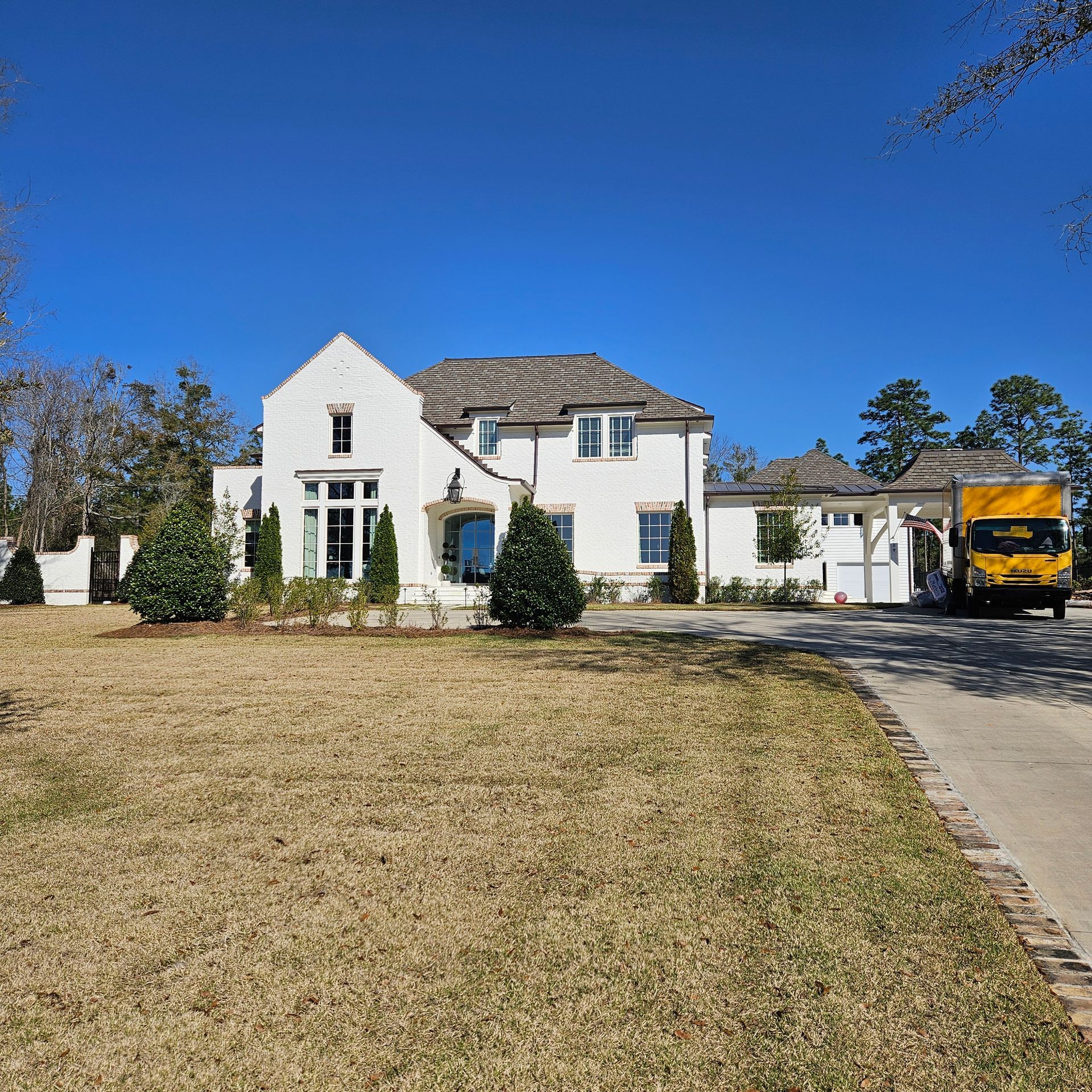 A white two-story house with a grey roof and a yellow delivery truck parked in the driveway on a sunny day.