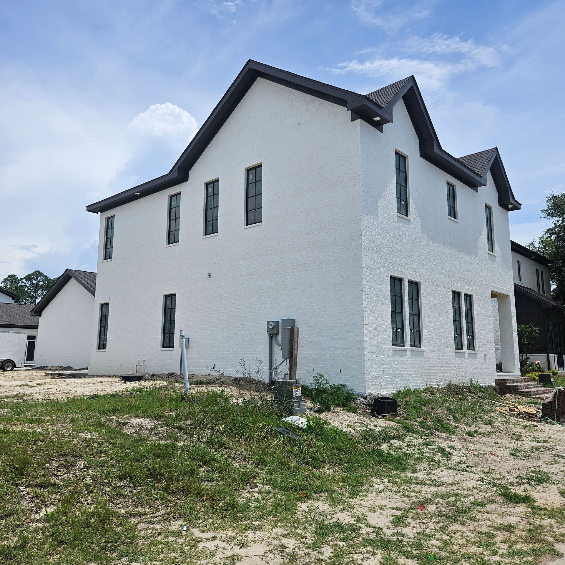 A two-story white brick house with a dark roof under a bright blue sky on a lot with patchy grass and sand.