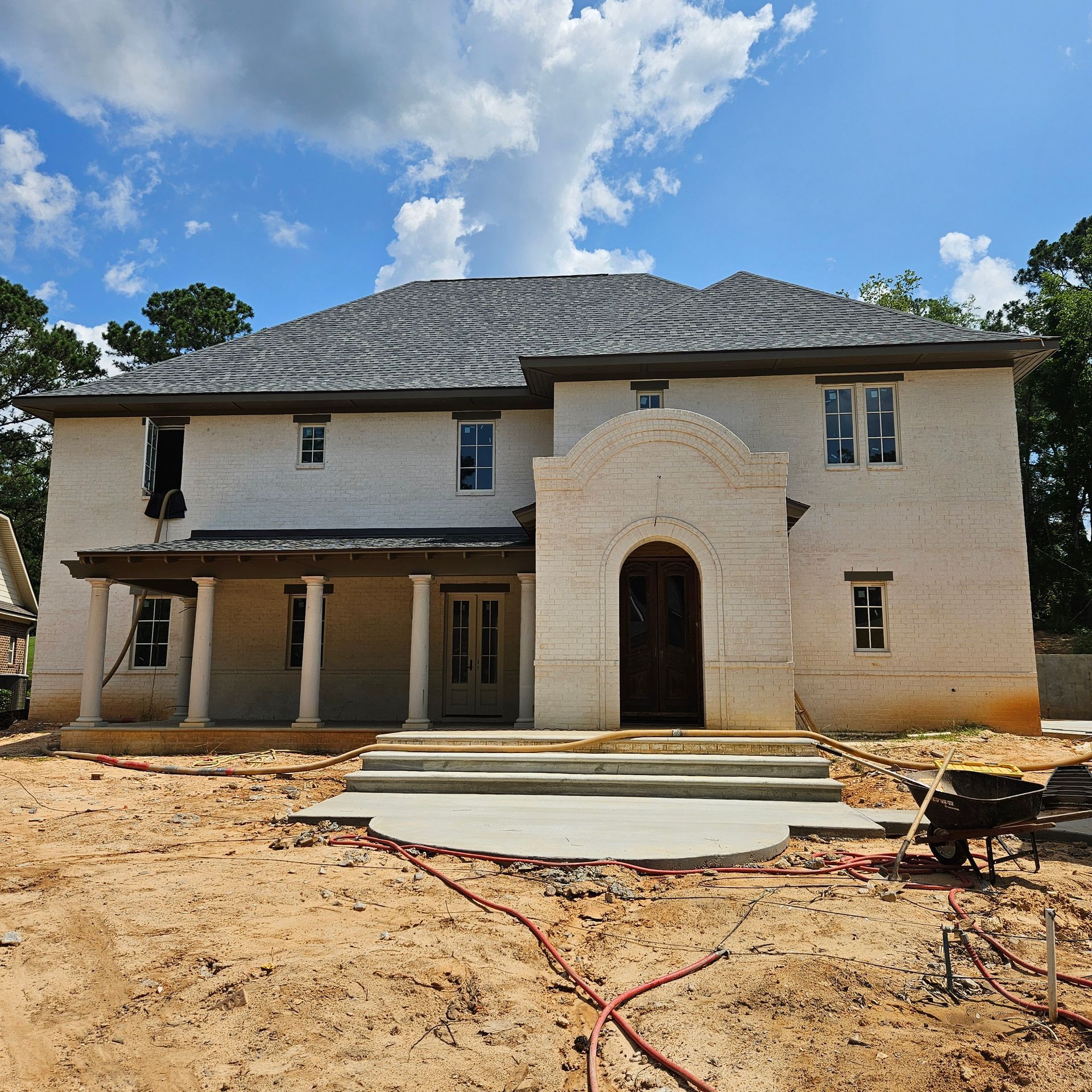 A partially constructed cream-colored house with a dark roof, arched entryway, and front porch on a dirt lot.