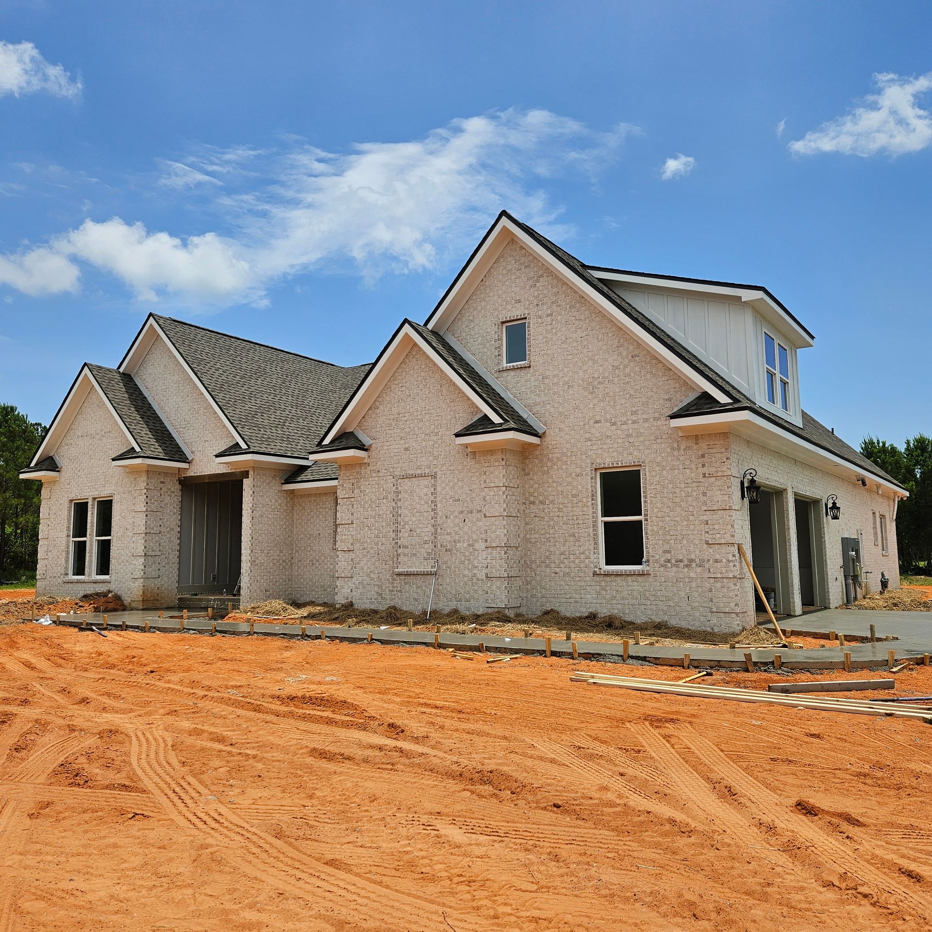 A newly constructed white brick house with a gray shingle roof sits on a dirt lot under a bright blue sky.