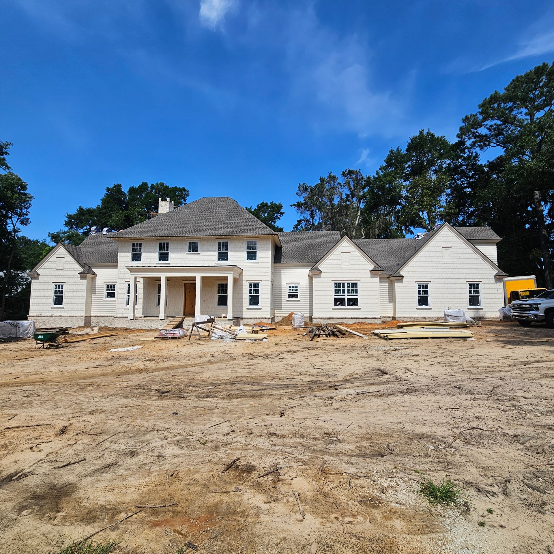 A large, single-family home under construction with light-colored siding and a dark shingled roof under a clear blue sky.