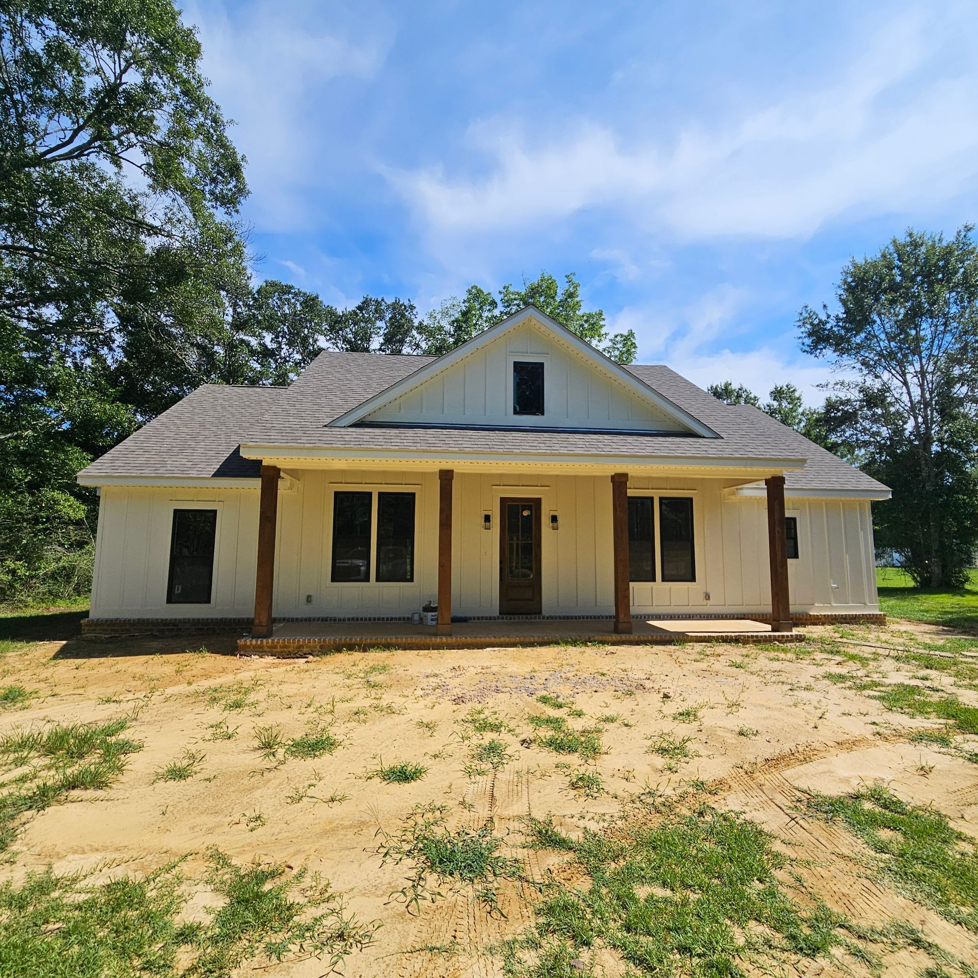 A newly built, white board-and-batten farmhouse with a covered porch and dark wood posts, set against a backdrop of trees.