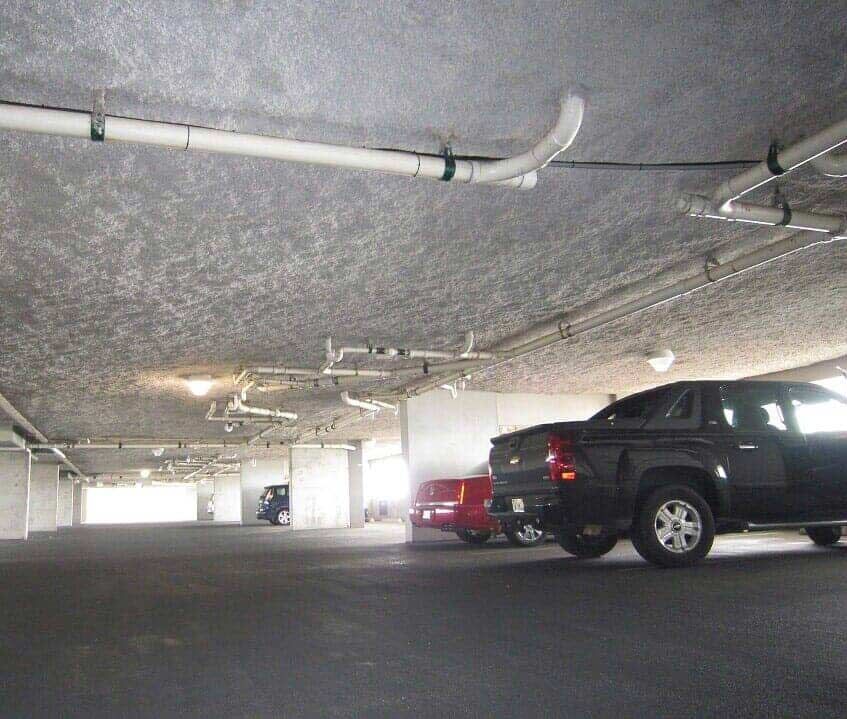 A parking garage with a low, textured concrete ceiling featuring pipes and lights, with a few cars parked in the stalls.