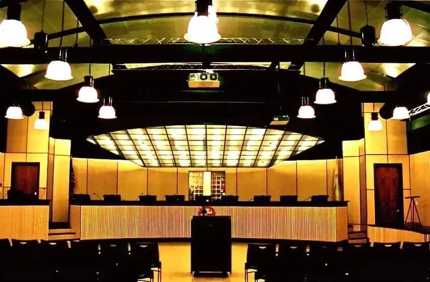 A courtroom featuring a long wooden dais, a central podium, rows of audience seating, and a large overhead grid light.