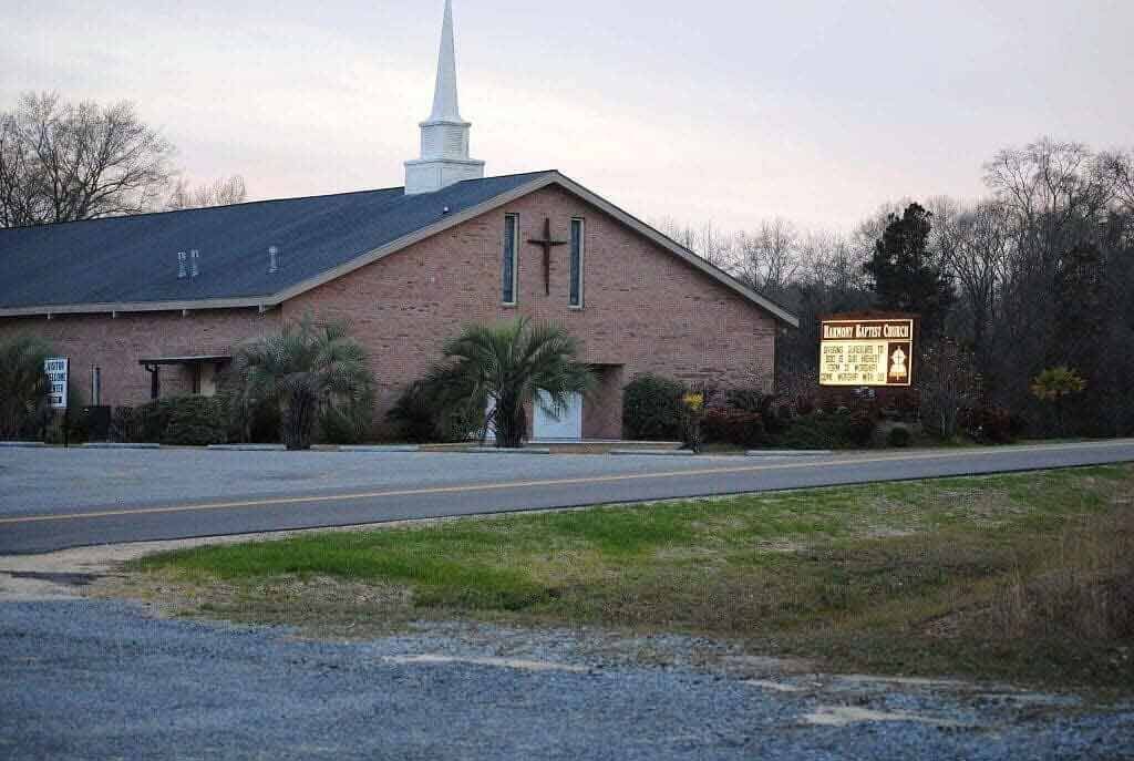 A tan brick church with a white steeple and a cross on the front, set against a tree-lined background at dusk.