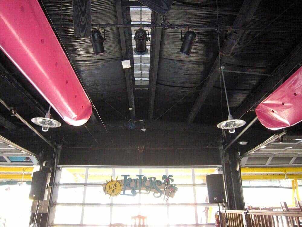 A restaurant interior with a dark ceiling, large pink ventilation ducts, pendant lights, and a glass garage door.