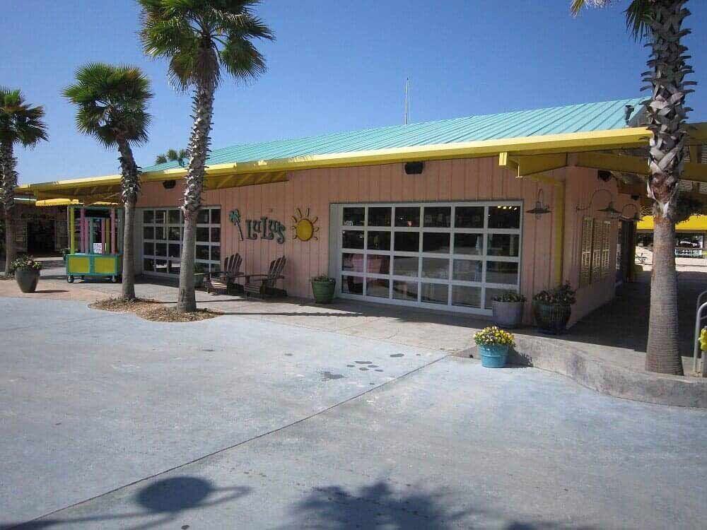 A pastel pink building with a teal metal roof, large glass garage-style doors, and palm trees under a bright blue sky.