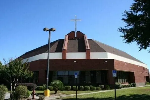 A modern red brick church with a large central cross on a dark shingled roof under a clear blue sky.
