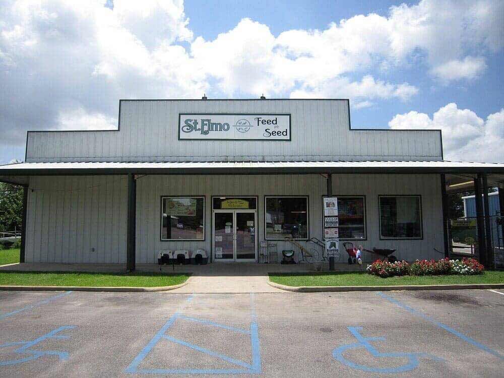 A white, single-story St. Elmo Feed & Seed store with a front porch, large windows, and a paved parking lot.