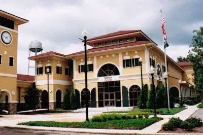 A pale yellow, two-story municipal building with a clock tower, water tower, and arched windows under a cloudy sky.