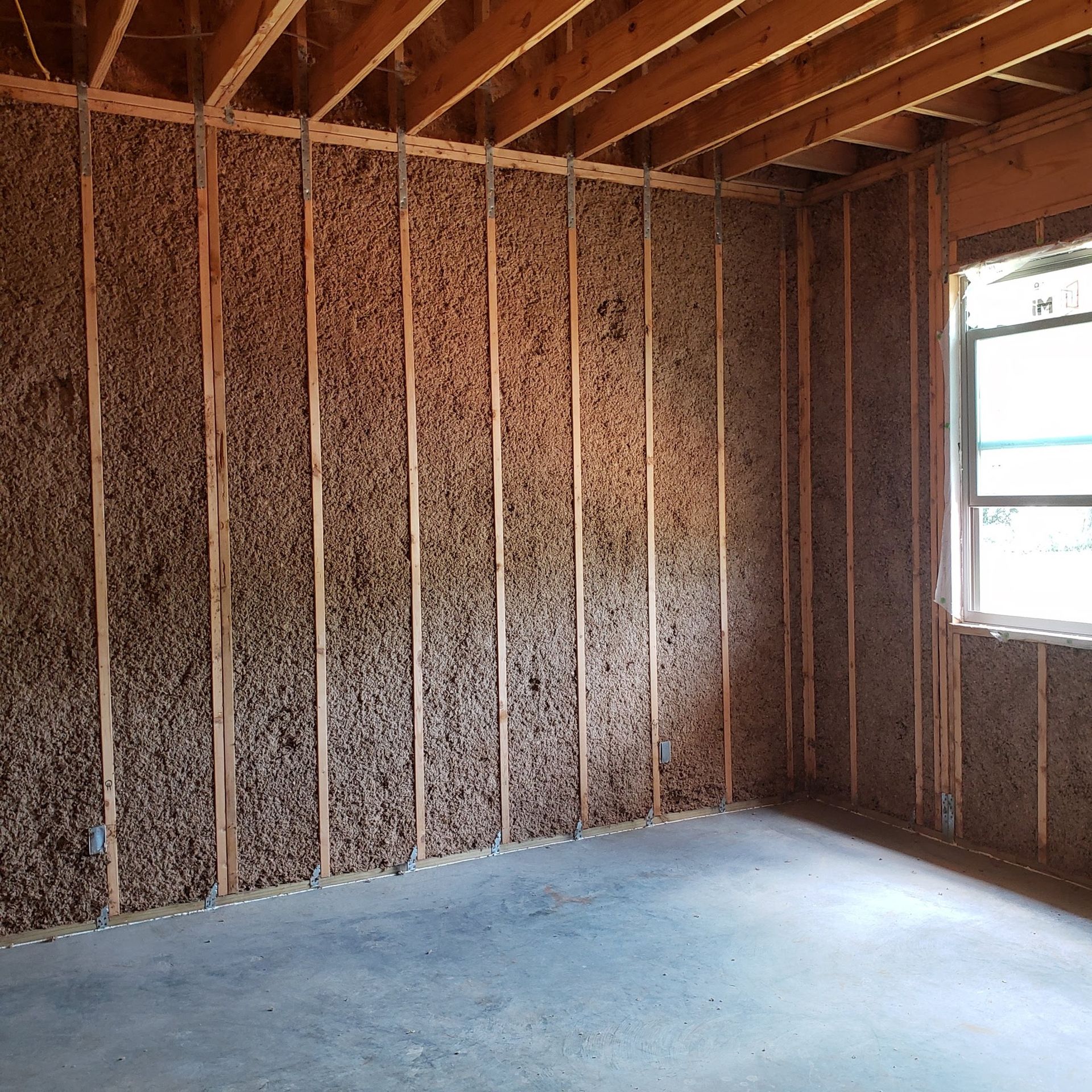 Unfinished interior room with exposed wooden wall studs, cellulose insulation, and a single window.