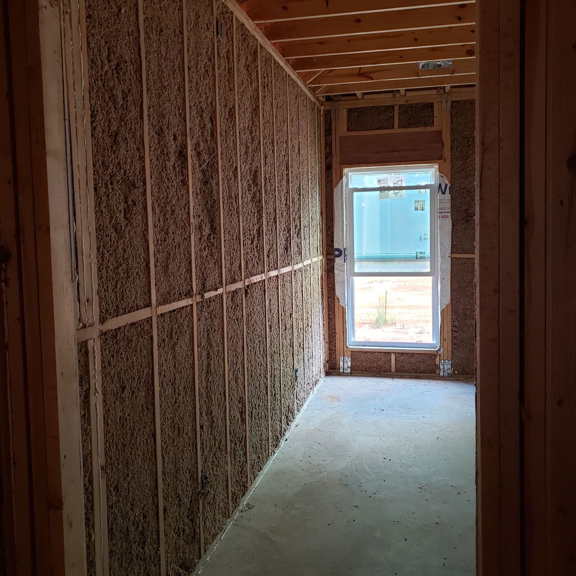 An unfinished hallway interior with brown fiber insulation installed between wall studs, leading to a framed window.