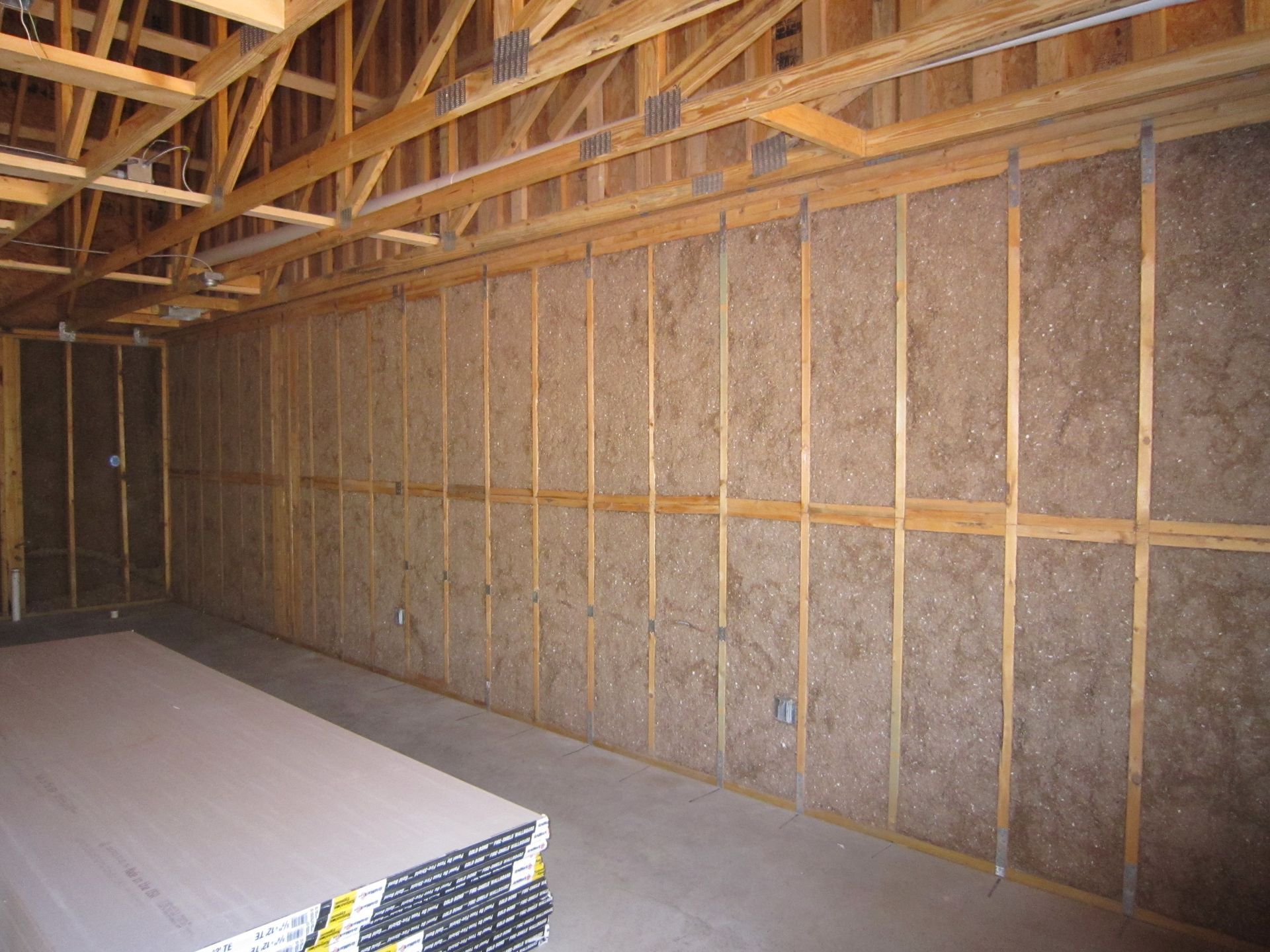 An unfinished room with exposed wooden wall studs filled with brown insulation, and a stack of drywall on the floor.