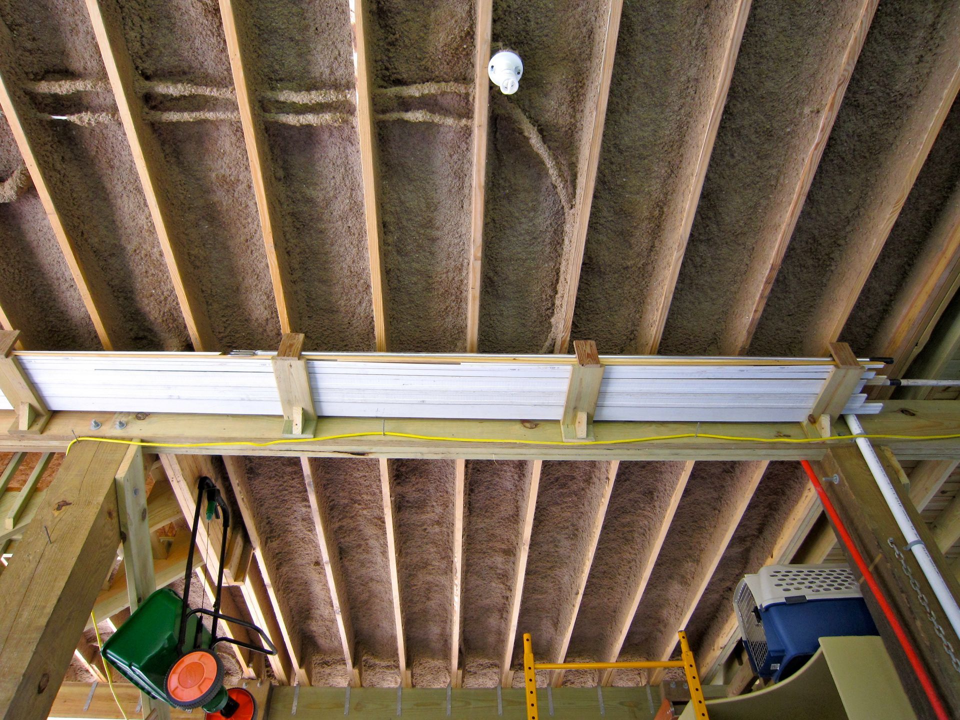An unfinished basement ceiling with exposed wooden joists, ceiling insulation, a utility light, and stacked trim boards.