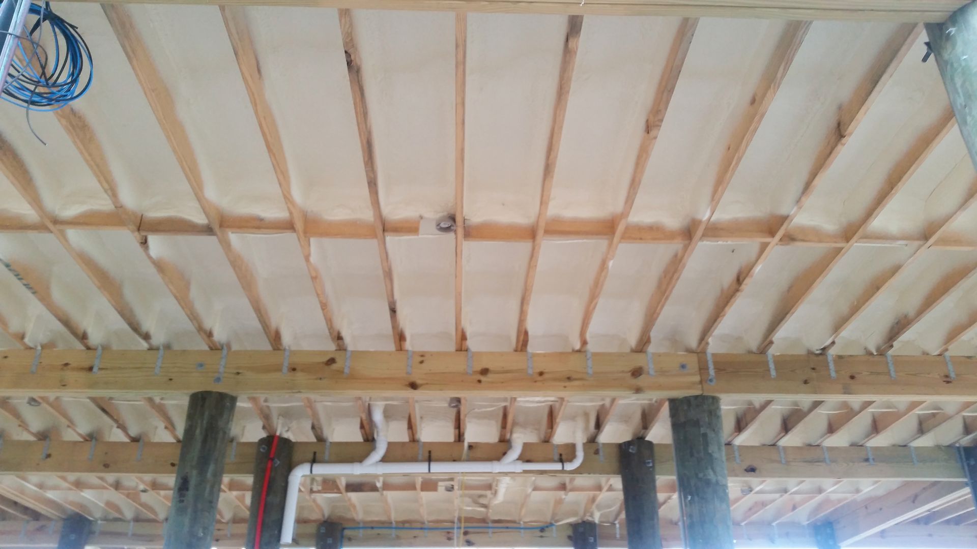 Insulated floor joists under a house structure with wooden beams and visible white plumbing pipes.