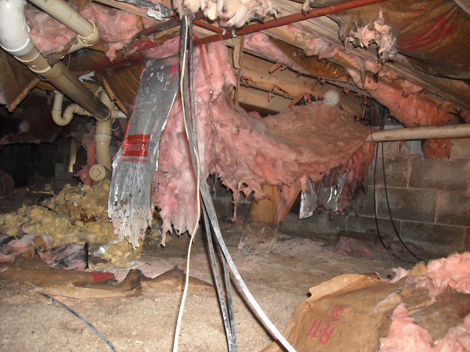 Damaged, sagging pink insulation and silver ducting hanging from the joists in a cluttered, unfinished crawl space.