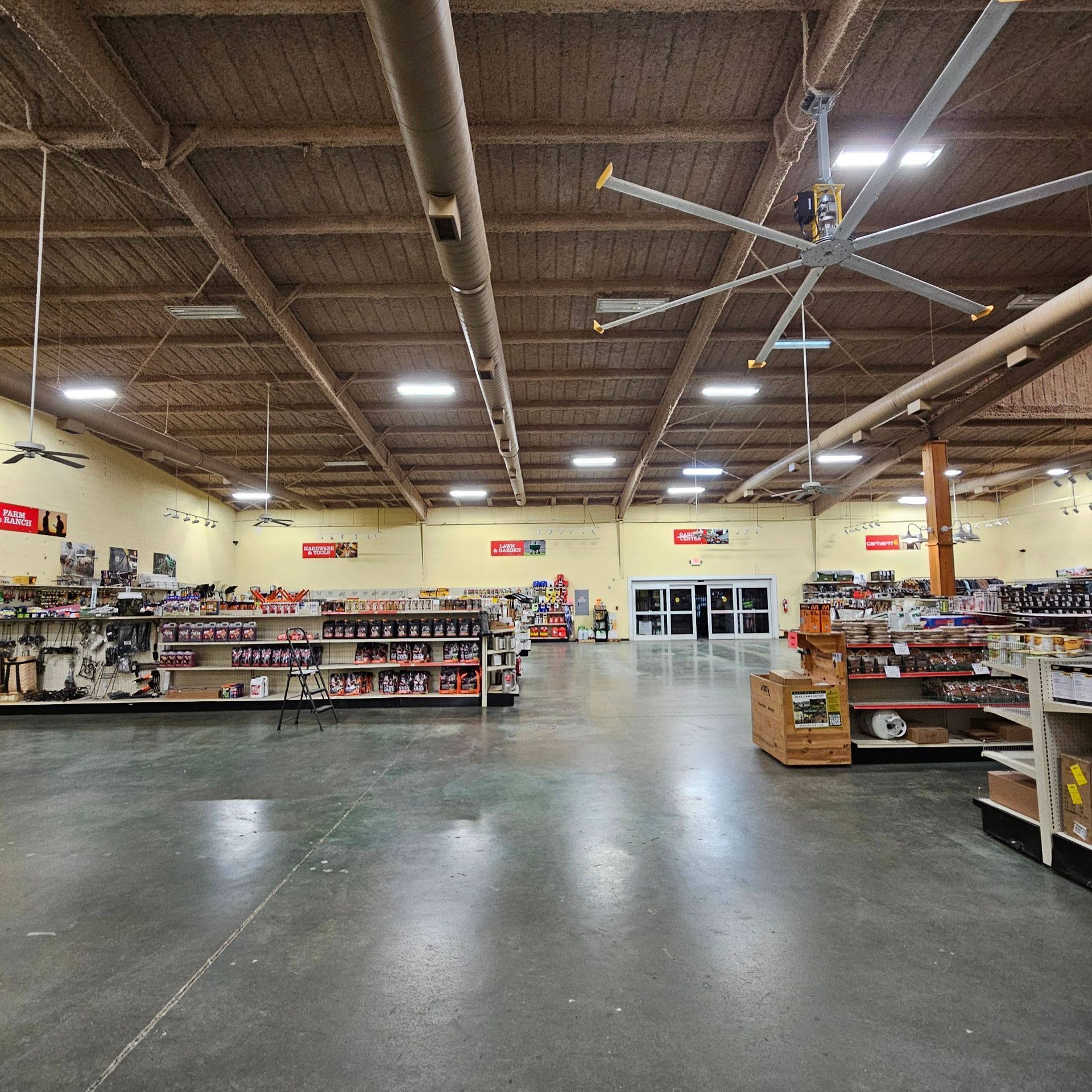 A large retail store with shelves of products, a high, open-joist ceiling, a large ceiling fan, and concrete flooring.