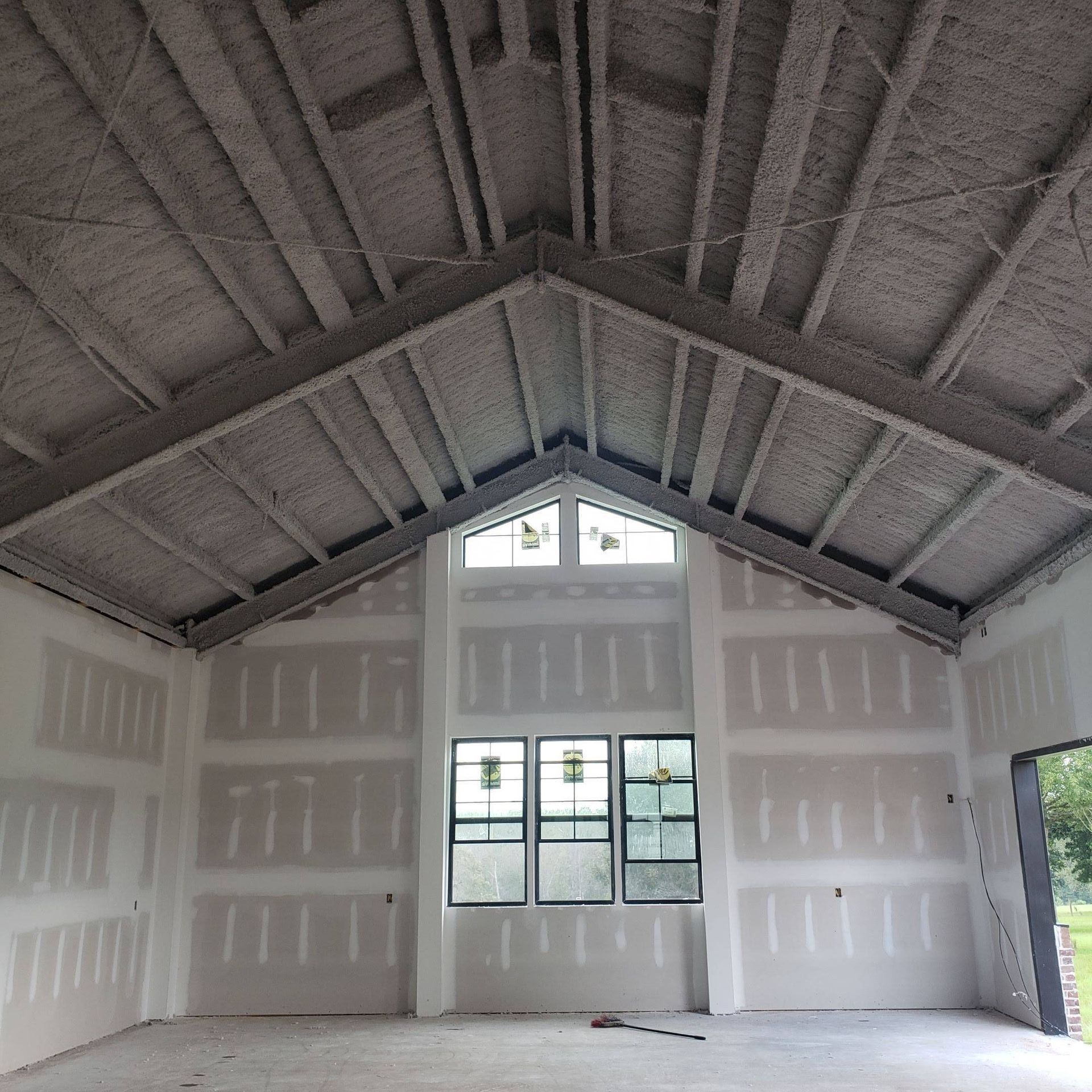 An interior view of a room under construction with unfinished drywall, exposed ceiling beams, and a large window.