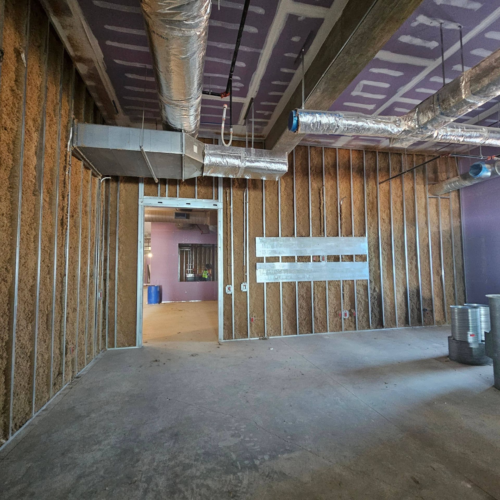 An interior view of a commercial construction site with metal wall studs, exposed ductwork, and purple ceiling panels.