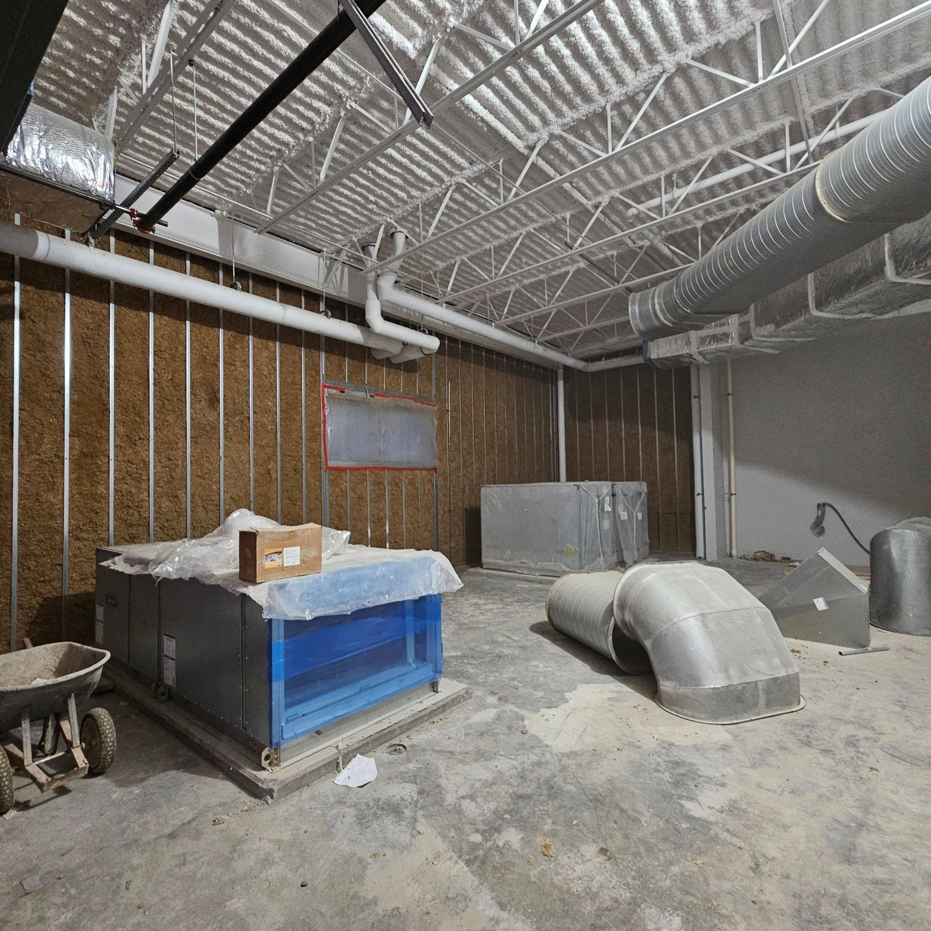 An industrial space under construction with exposed metal studs, ventilation ducts, and HVAC equipment on a concrete floor.